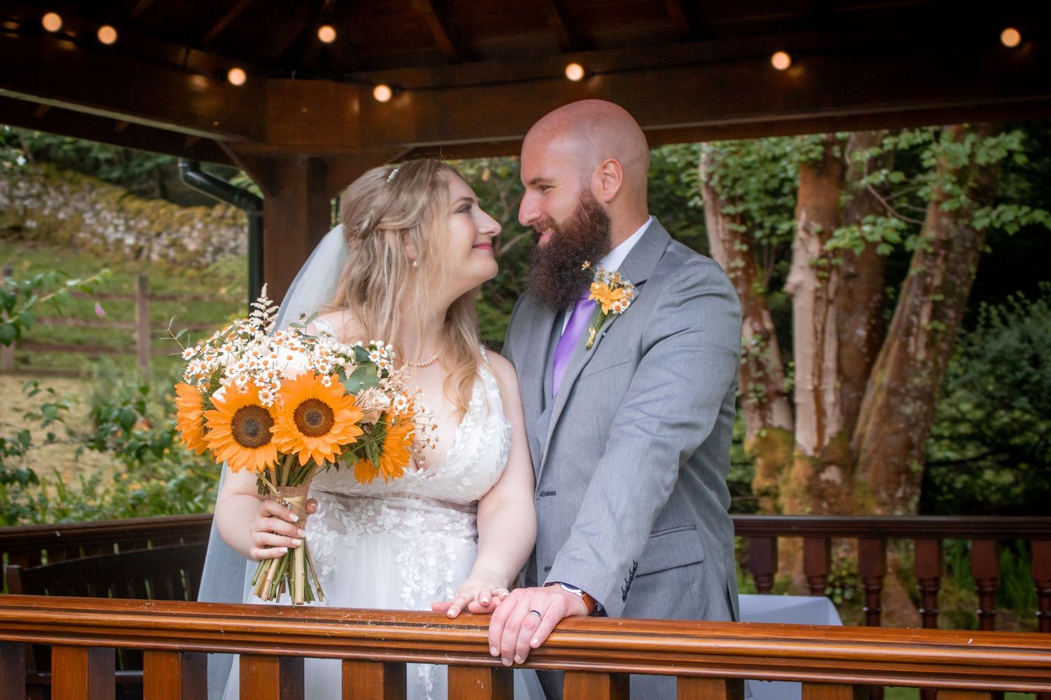 Bride and groom smile at each other on a wooden deck, bride holding vibrant sunflower bouquet.