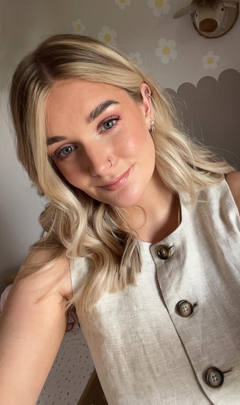 Young blonde woman taking a selfie wearing a beige linen vest top in a floral decorated room.