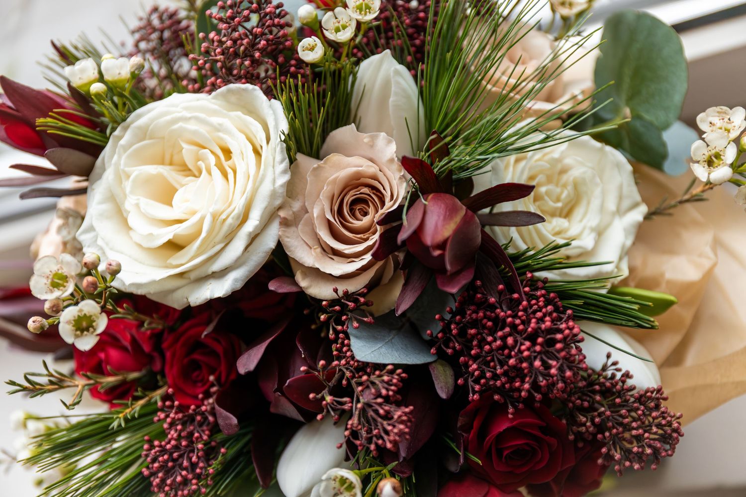 Elegant bridal bouquet featuring cream roses, dark red blooms, pine sprigs, and berry accents for a winter wedding.