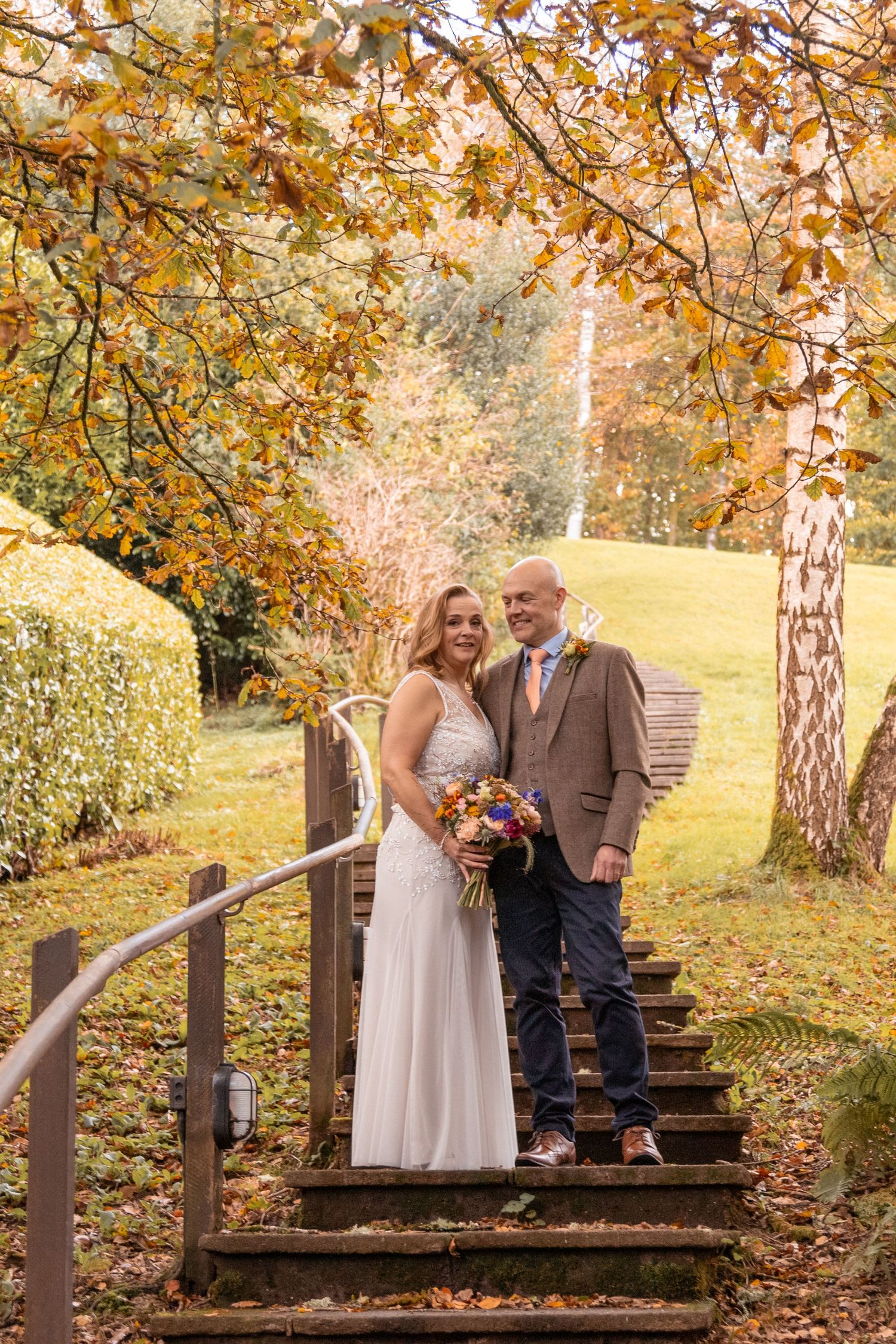 Smiling bride and groom posing on wooden steps surrounded by golden autumn foliage.