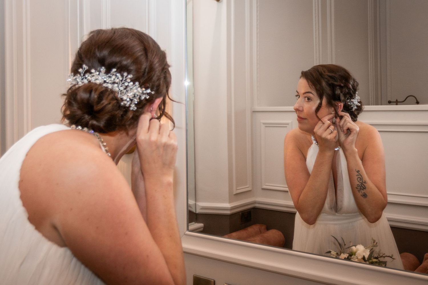 Bride in white gown adjusting earring while looking in mirror, wearing elegant updo with pearl hair accessory.
