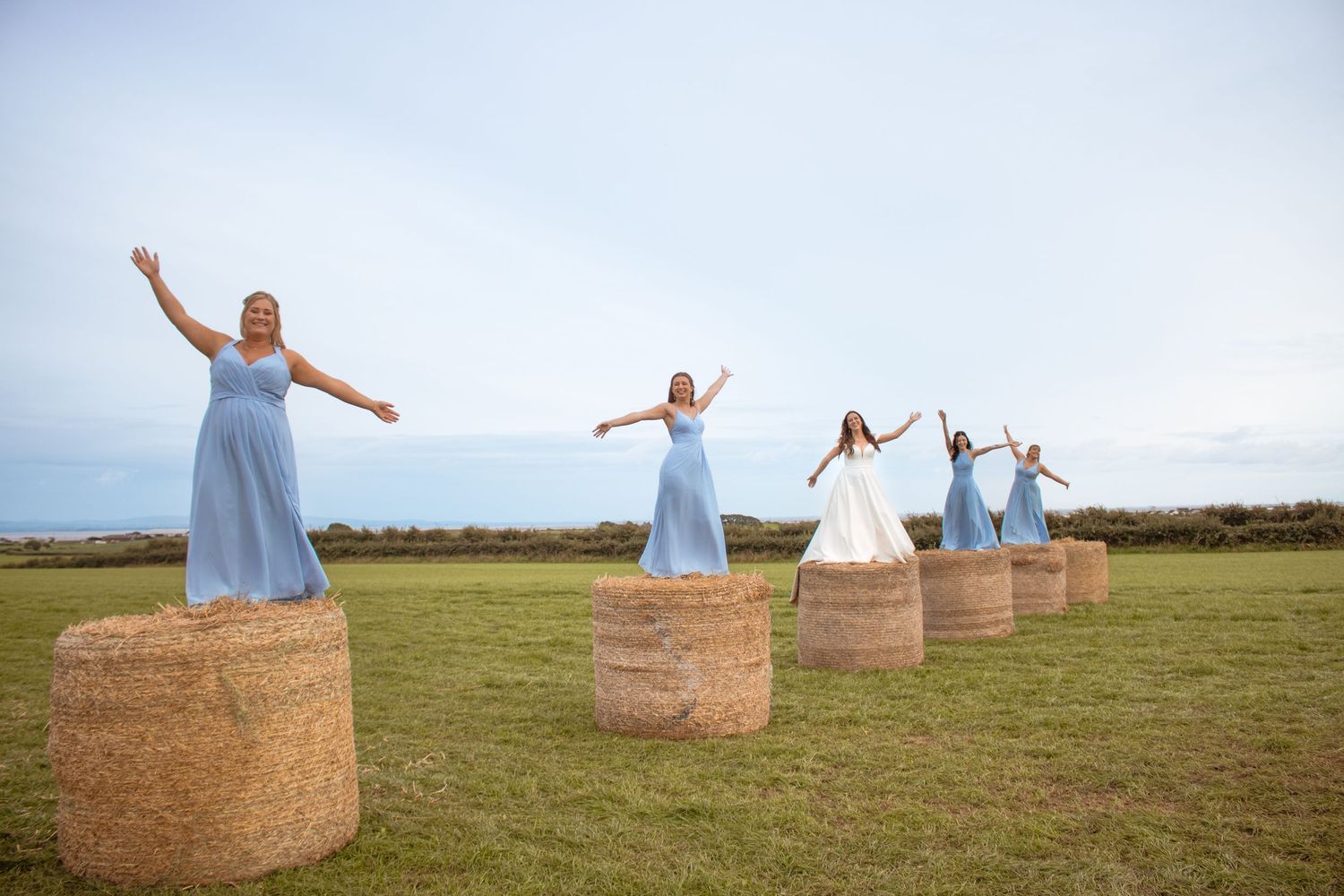 Bride and bridesmaids in blue dresses strike joyful poses standing atop hay bales in open field.