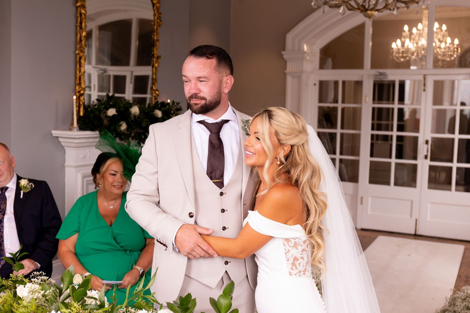 Bride and groom exchange vows at elegant wedding ceremony, bride in white lace gown with veil, groom in beige suit.