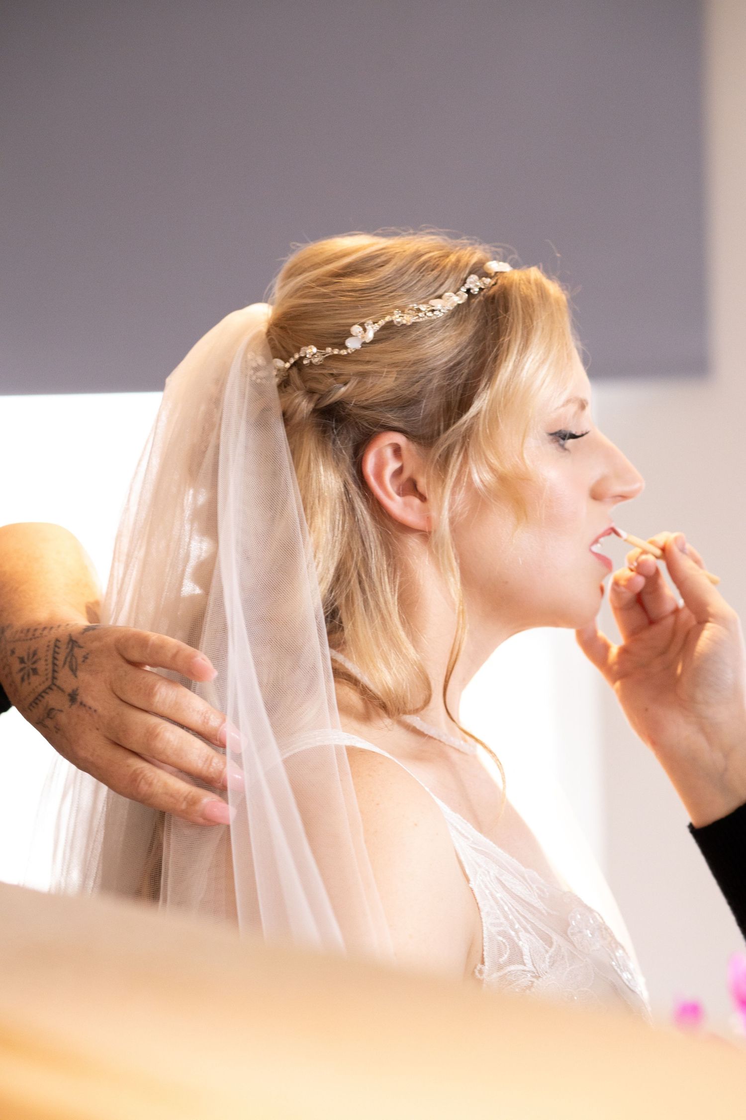 Blonde bride in white dress and floral hair vine veil gets makeup applied before her wedding ceremony.