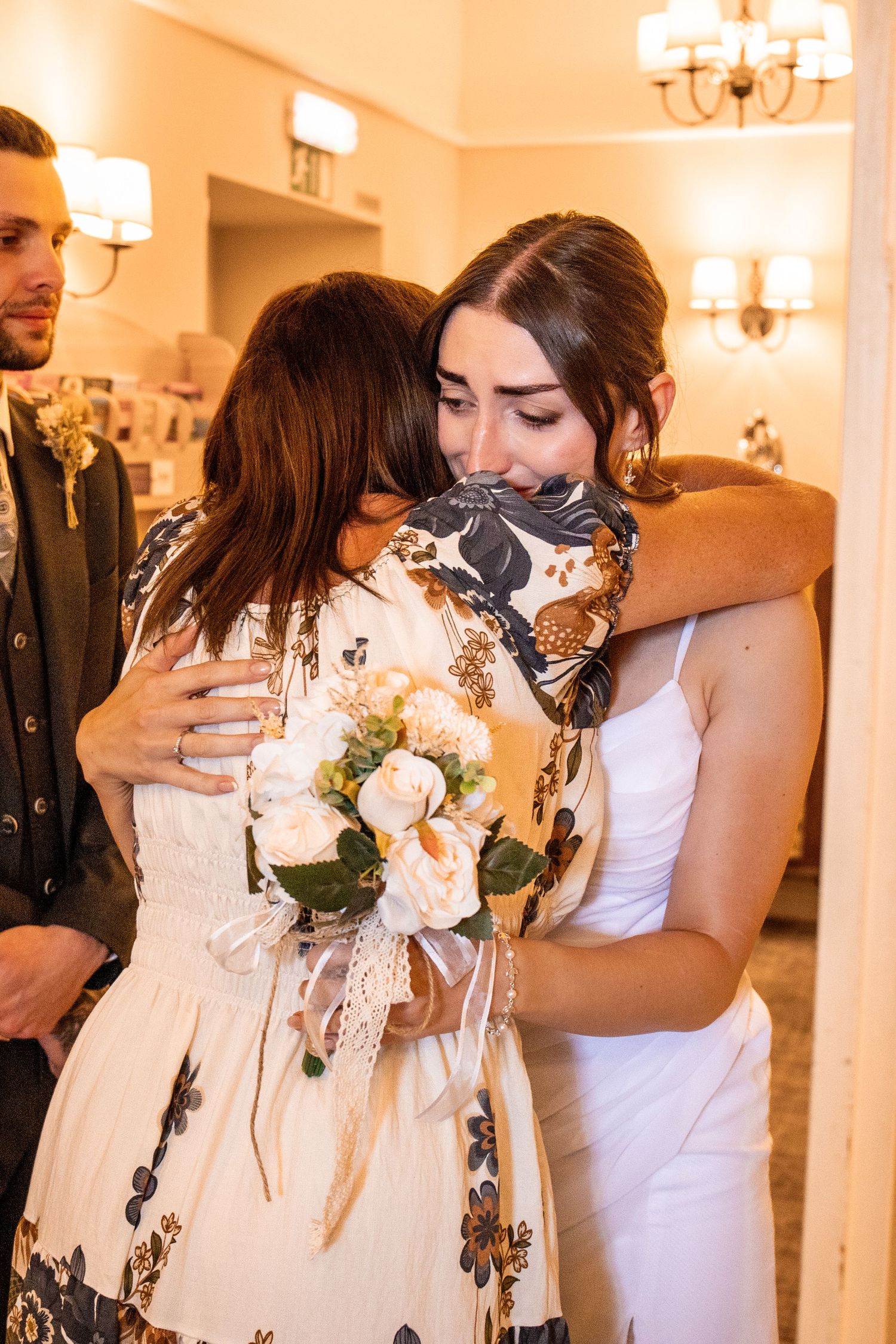 Two women sharing an emotional hug at a wedding, one in white bridal gown holding a bouquet of white roses.