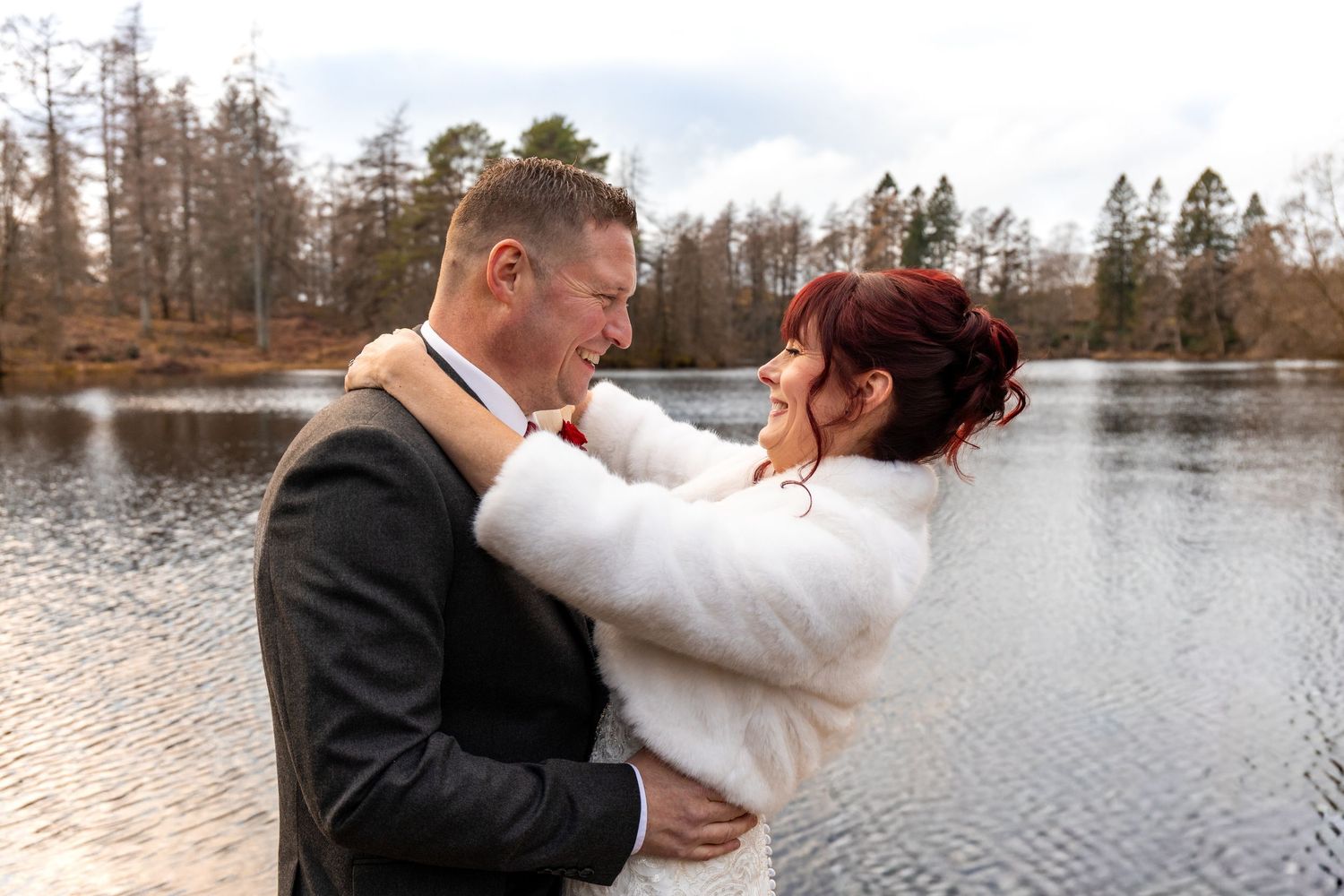 Bride in white fur wrap and groom share a joyful embrace outdoors by a lake with autumn trees.