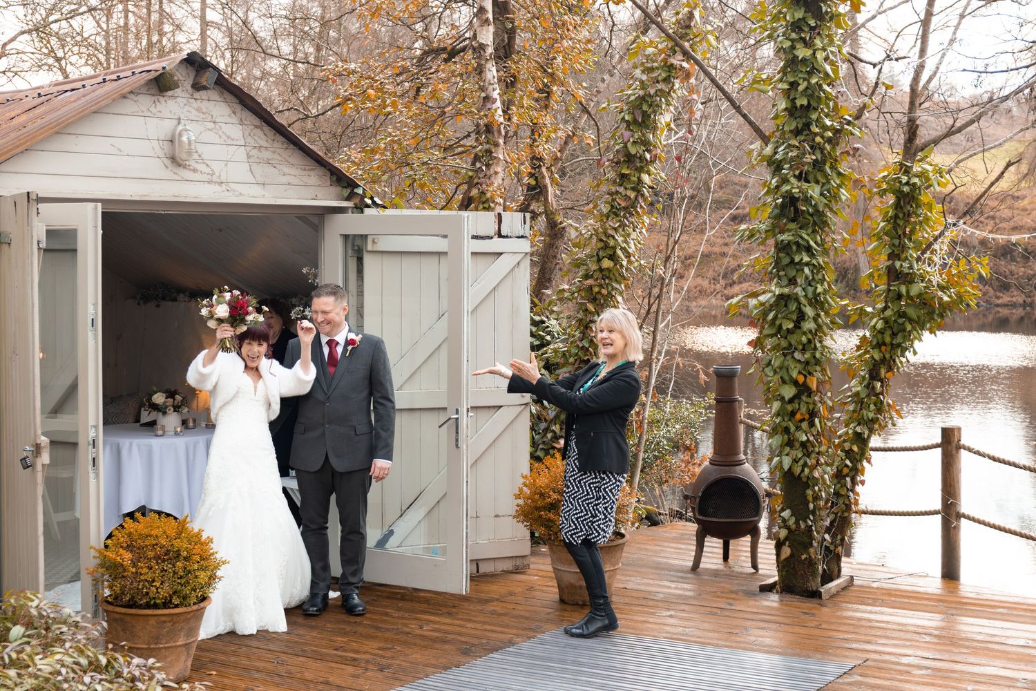 Bride and groom posing with officiant outside a rustic wooden chapel by a pond in autumn.