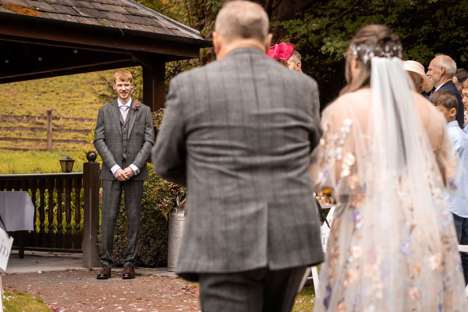 Groom smiling as bride walks down the aisle at an outdoor wedding ceremony surrounded by autumn foliage.