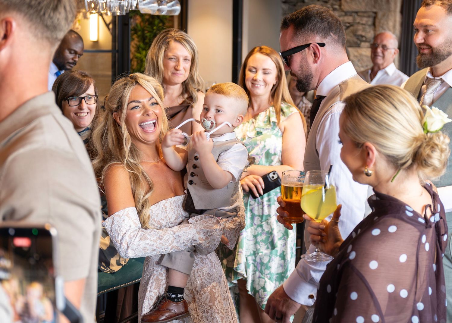 Bride laughing and celebrating with wedding guests indoors, holding drinks at a lively wedding reception.