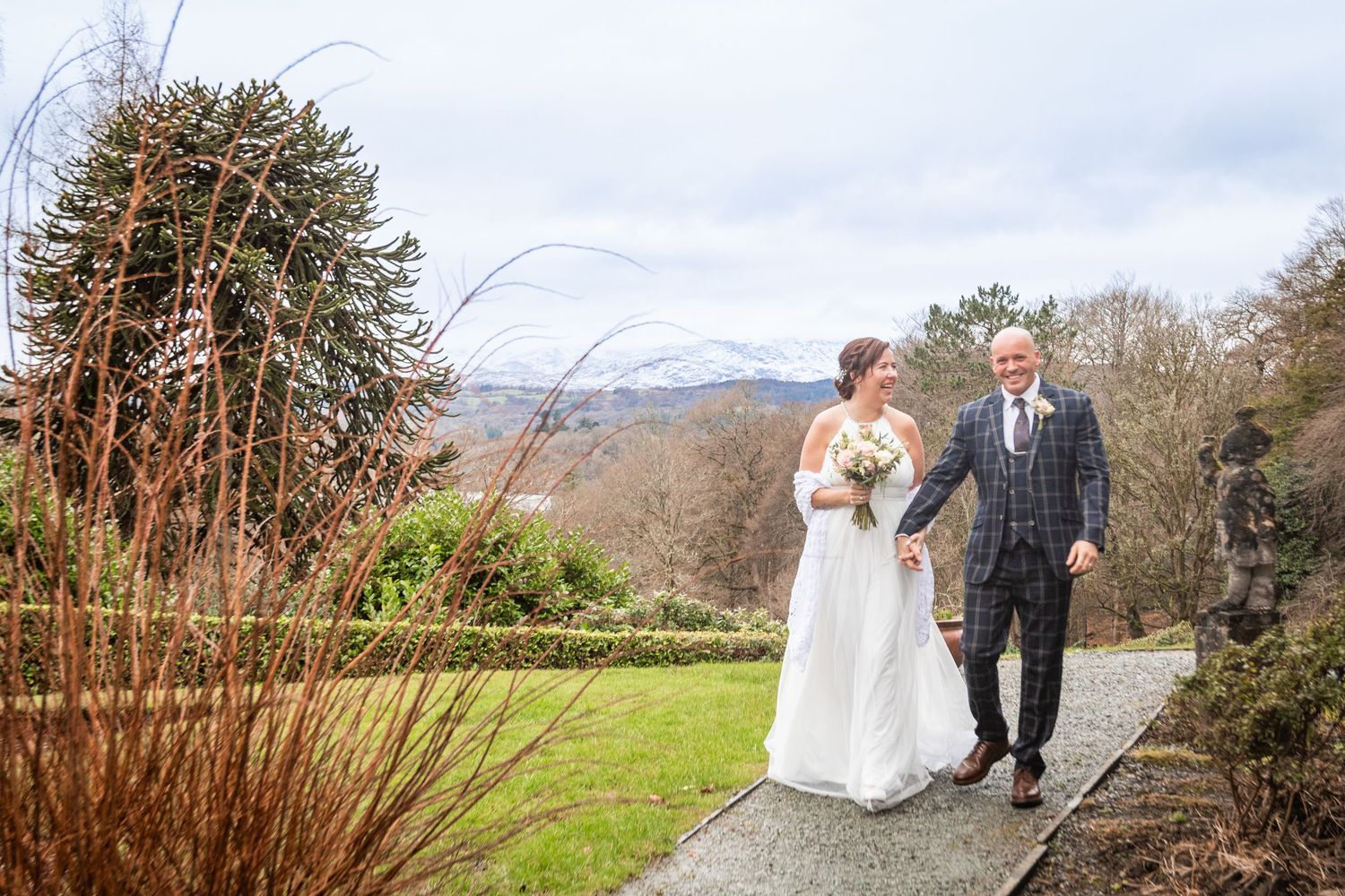 Bride and groom walk hand in hand along a garden path with scenic mountain views on their wedding day.