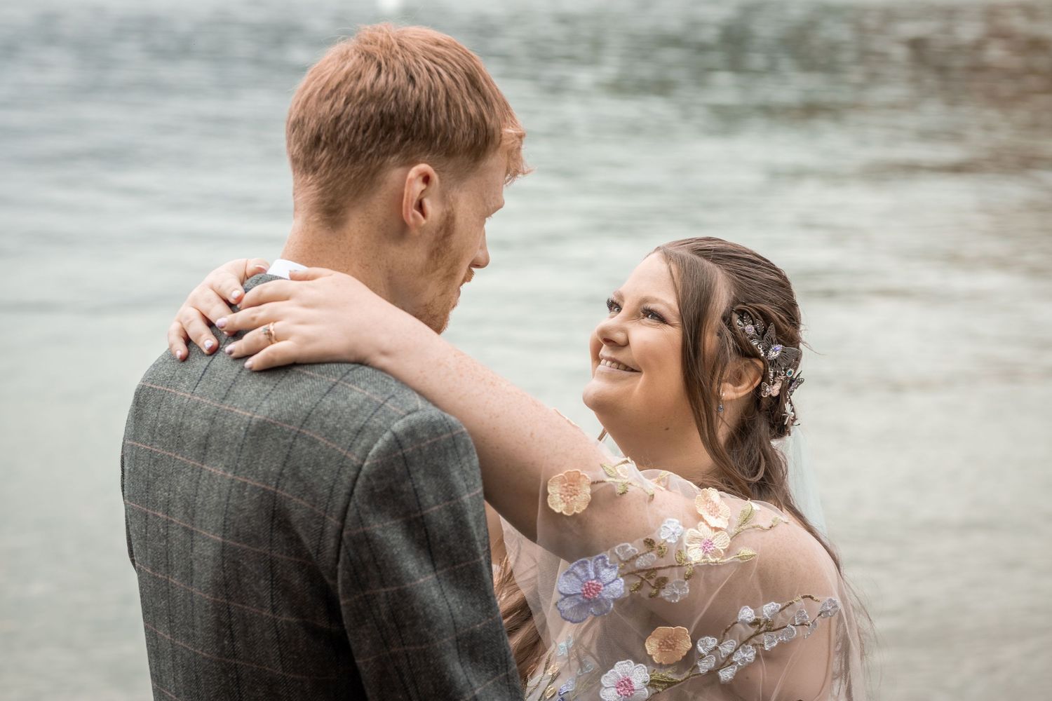 A smiling bride in a floral gown embraces a groom in a grey tweed suit near a shimmering lake backdrop.