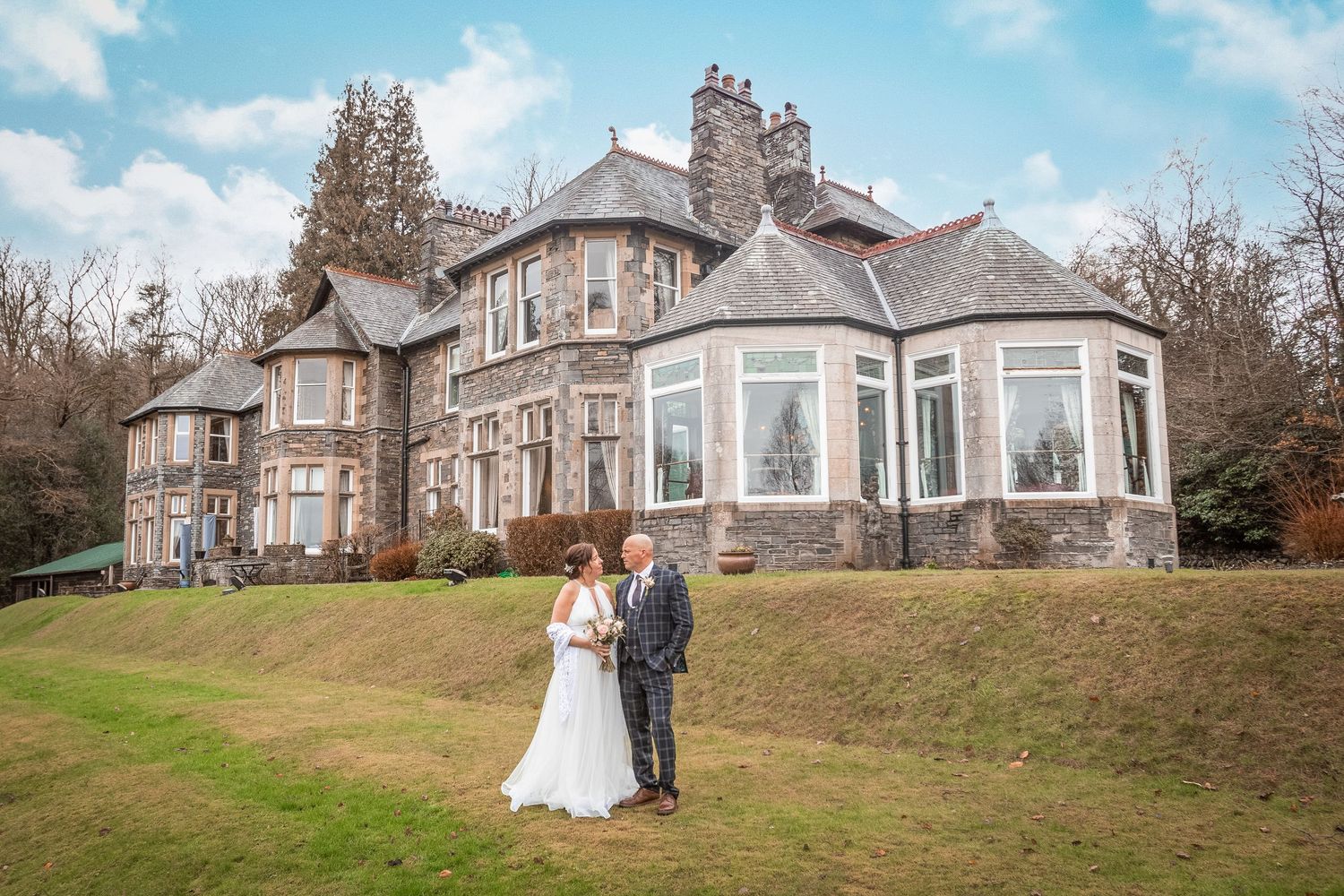 Bride and groom standing outside grand stone manor house on green lawn.