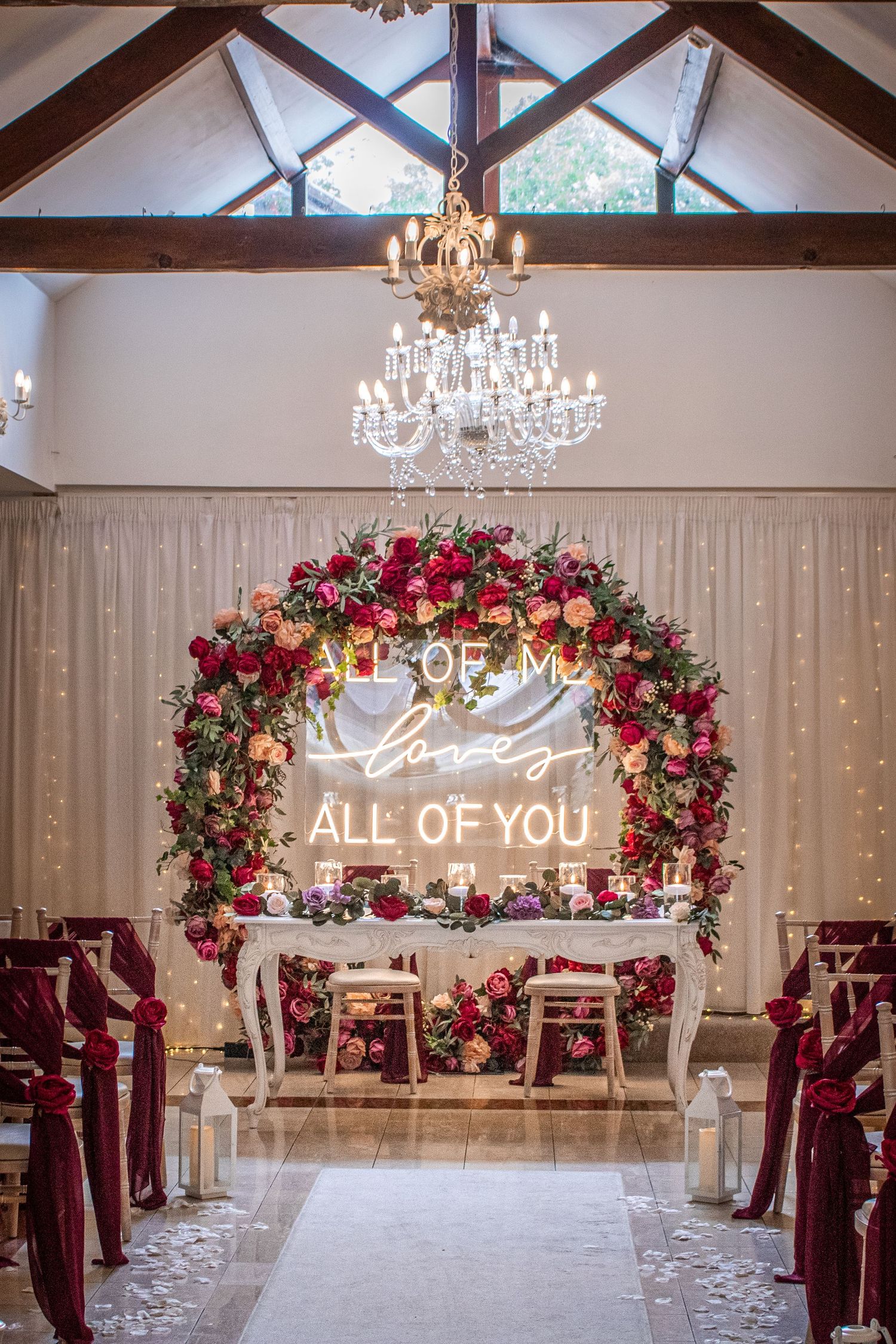 Elegant wedding ceremony venue with a floral arch, neon sign reading All of Me Loves All of You.
