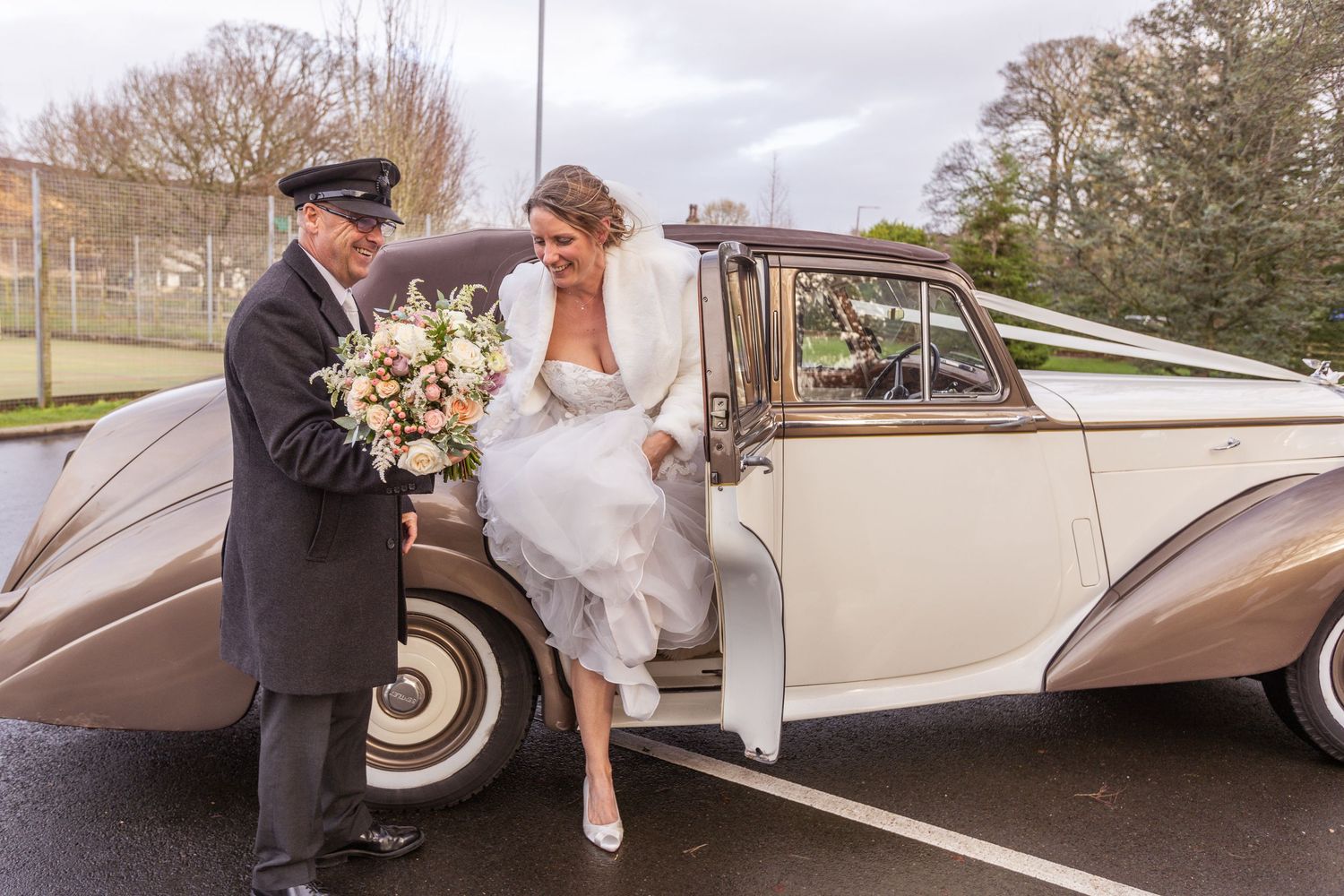 Bride stepping out of a vintage classic wedding car assisted by a chauffeur in a parking lot.