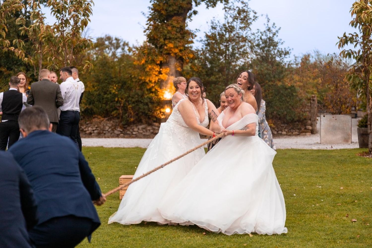 Two brides in white gowns laughing while playing tug of war on a lawn at an outdoor wedding reception.