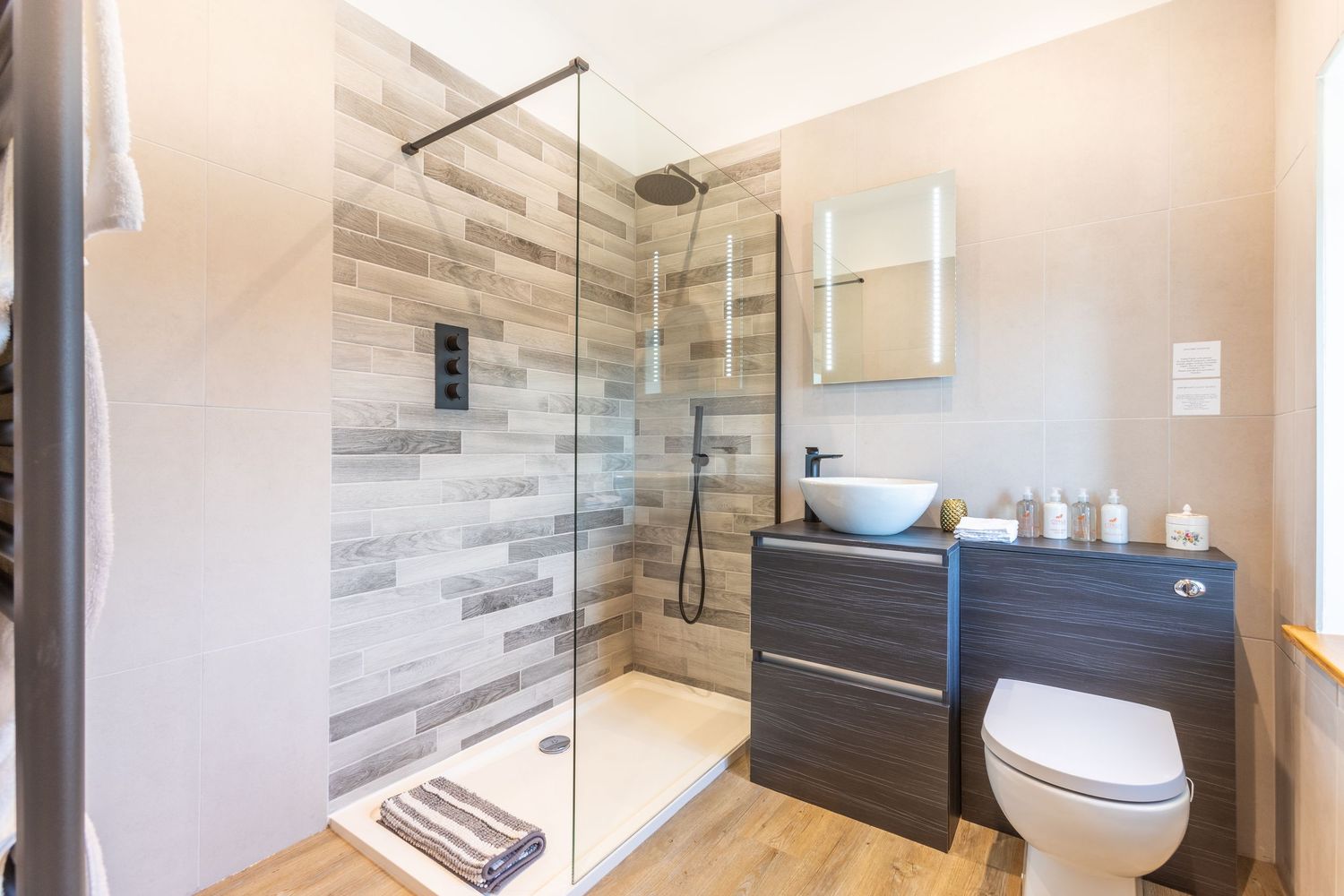 Modern en-suite bathroom with walk-in glass shower, wood-effect tiles, vessel sink, and wall-mounted toilet.