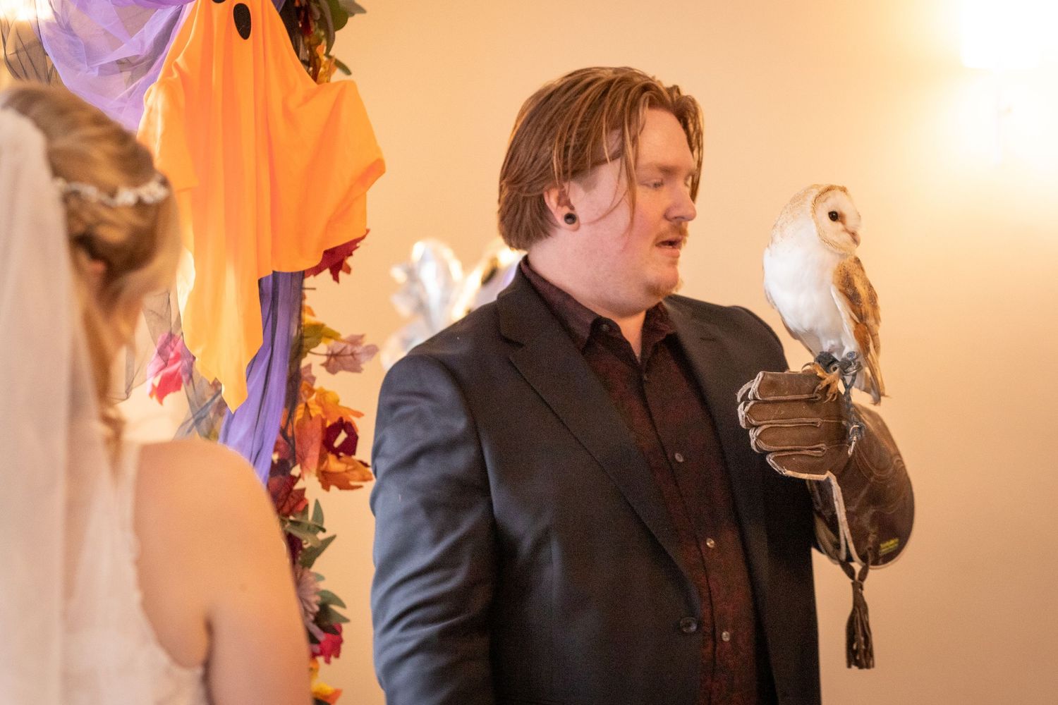 Man in dark suit holds a barn owl on a gloved hand at a colorful indoor event.