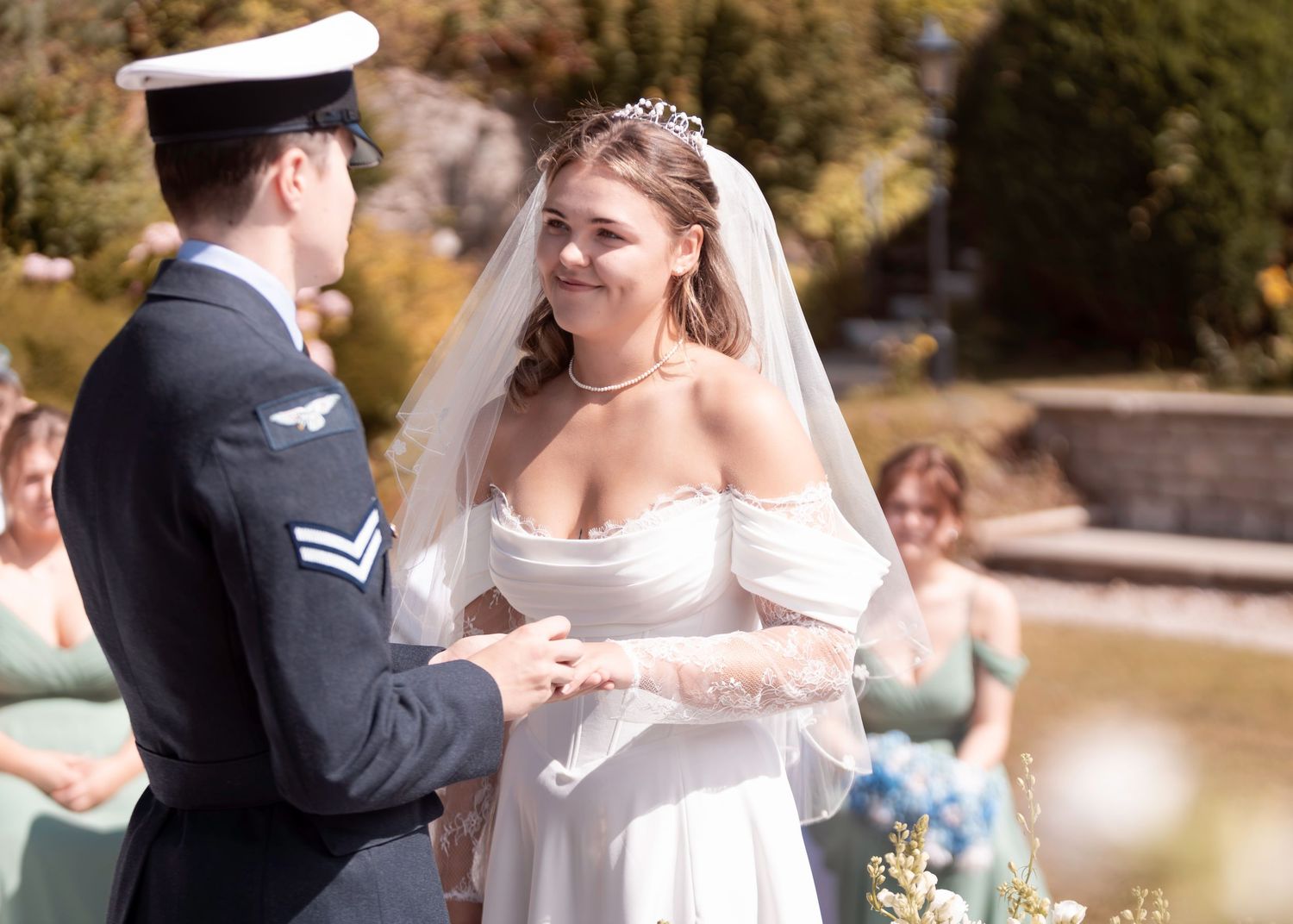 Bride in white off-shoulder gown and veil exchanges rings with groom in military uniform at outdoor wedding ceremony.