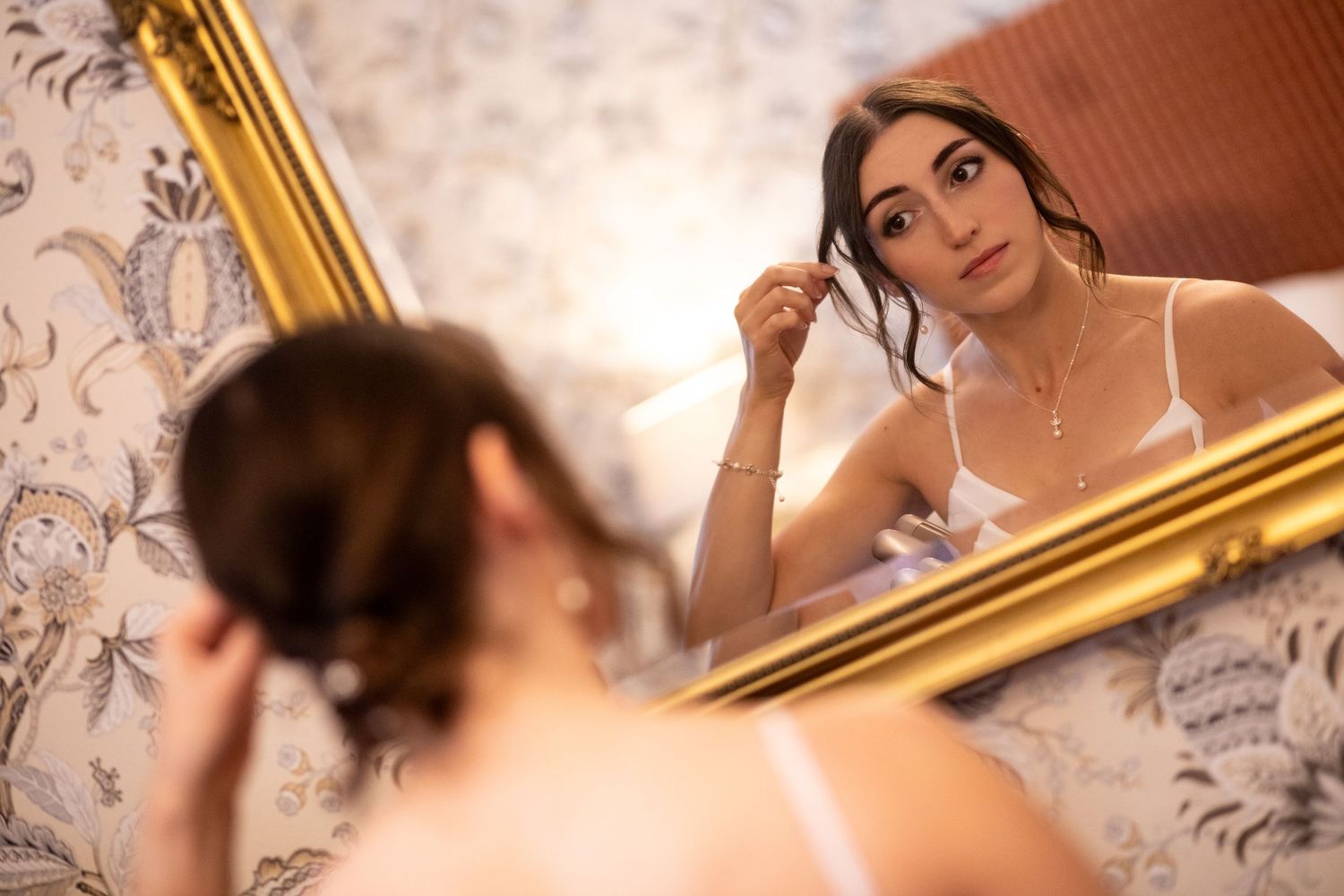Bride adjusting earring while gazing into ornate gold-framed mirror in elegantly wallpapered room.
