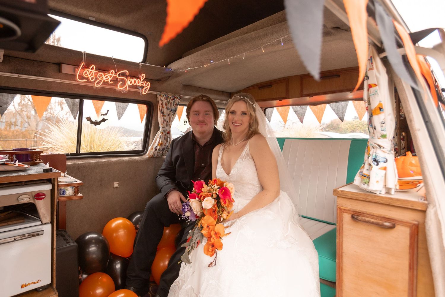 Bride and groom smiling inside a decorated vintage camper van with Halloween bunting and orange balloons.