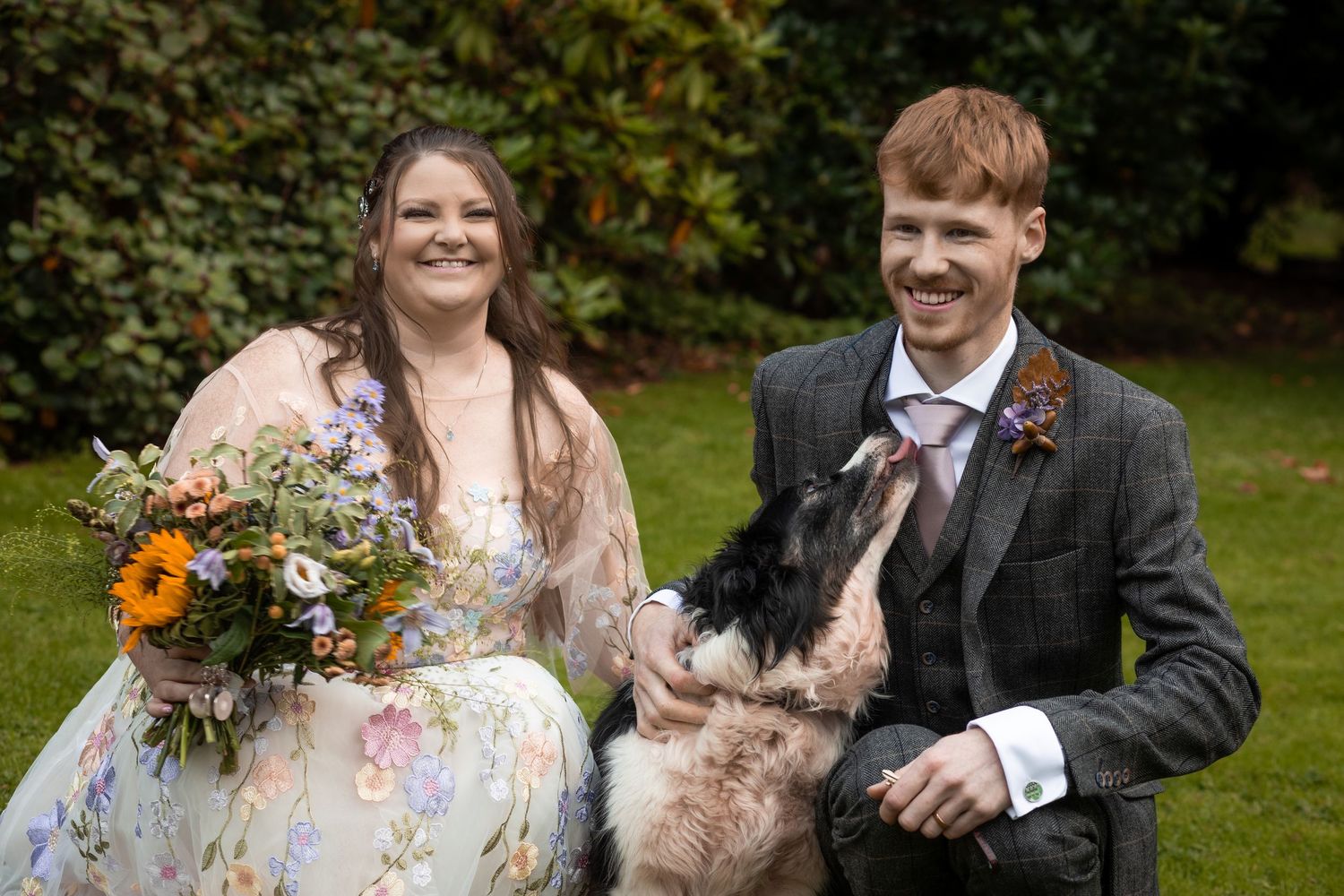 Happy newlyweds smile with their dog at an outdoor wedding ceremony on a lush green lawn.