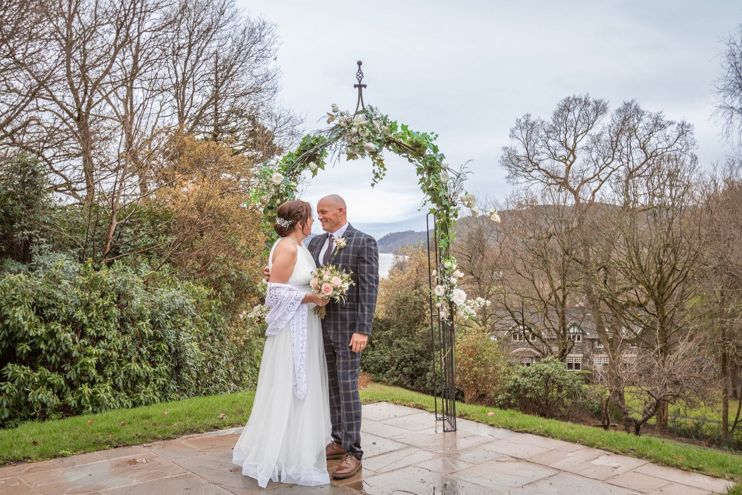 Bride and groom standing together under a decorated floral arch on a stone terrace with garden views.