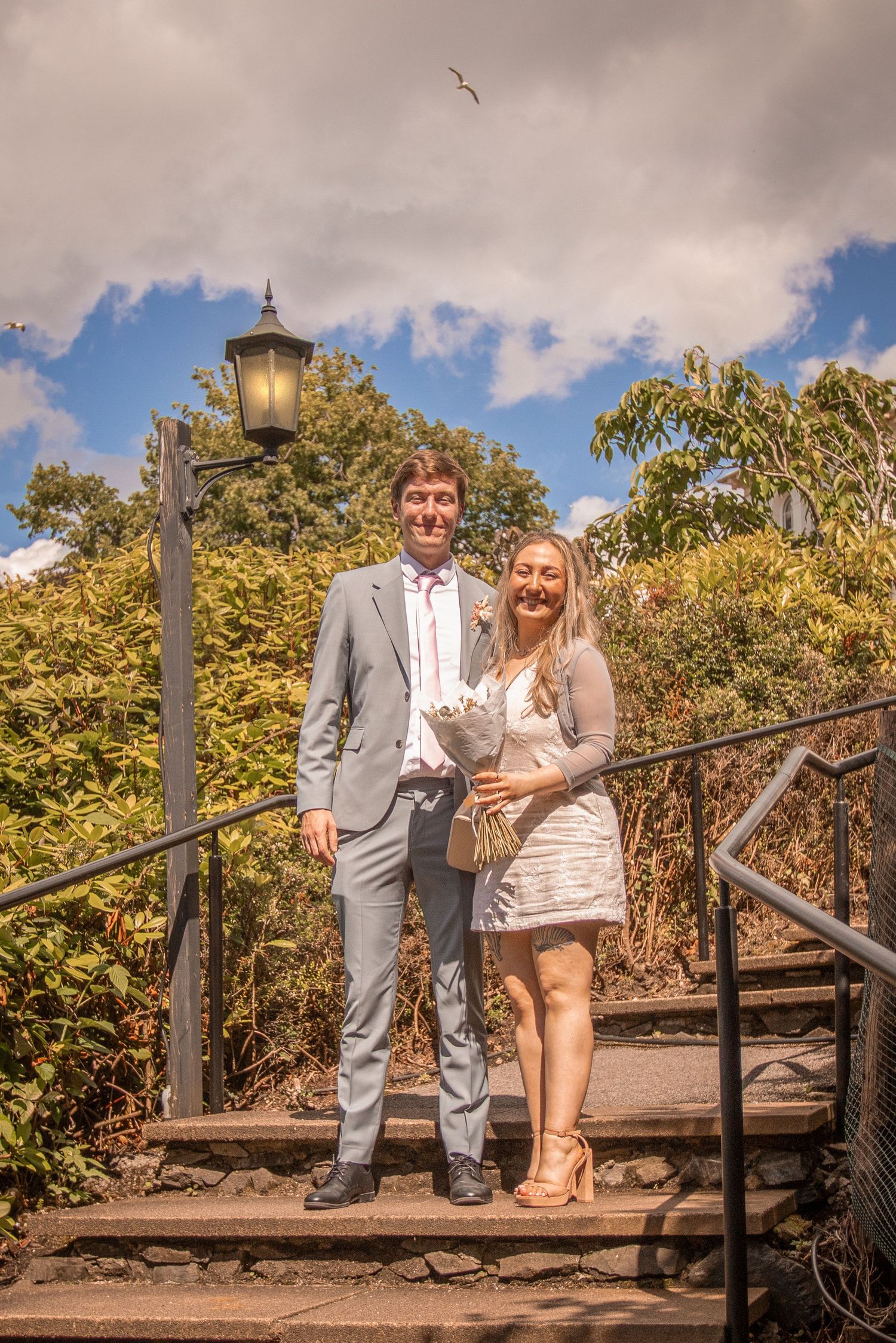 A smiling bride and groom pose together on outdoor steps surrounded by greenery on their wedding day.