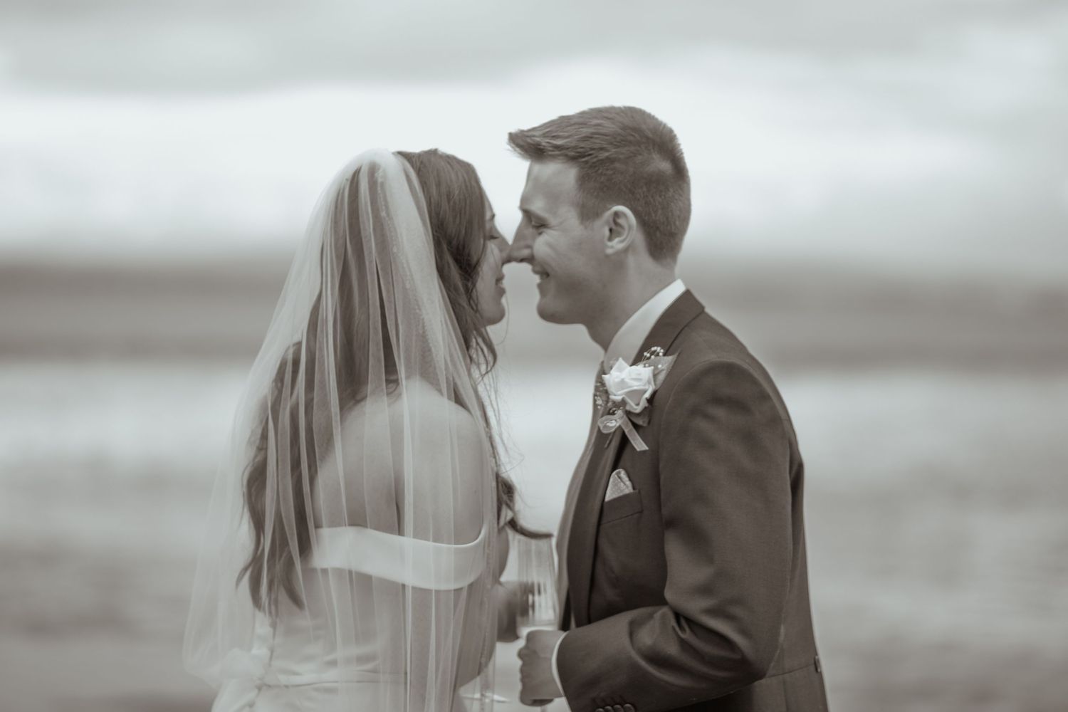 Black and white romantic beach wedding photo of bride and groom almost kissing near the ocean.