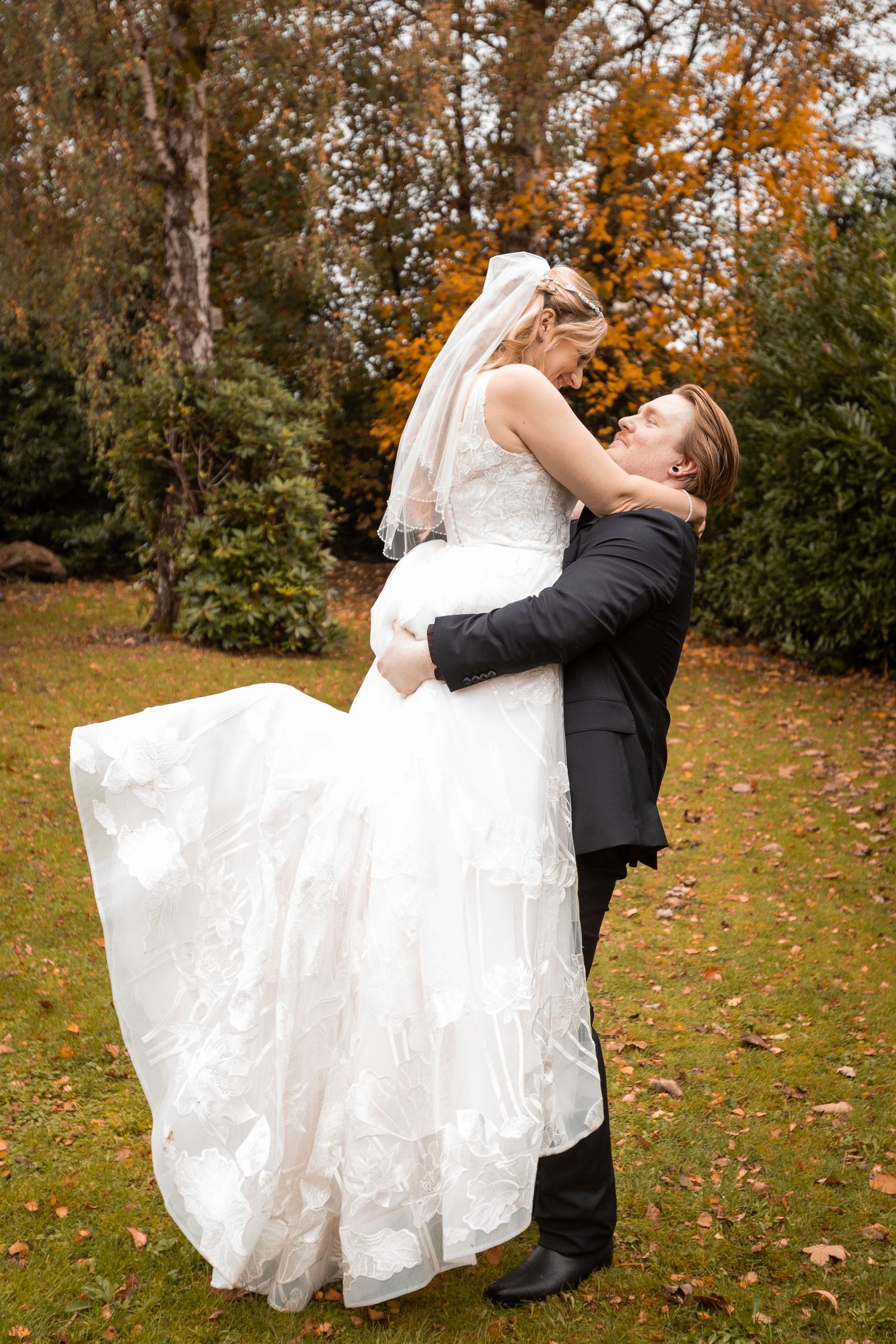 Groom lifting bride in white wedding dress outdoors among autumn foliage trees.