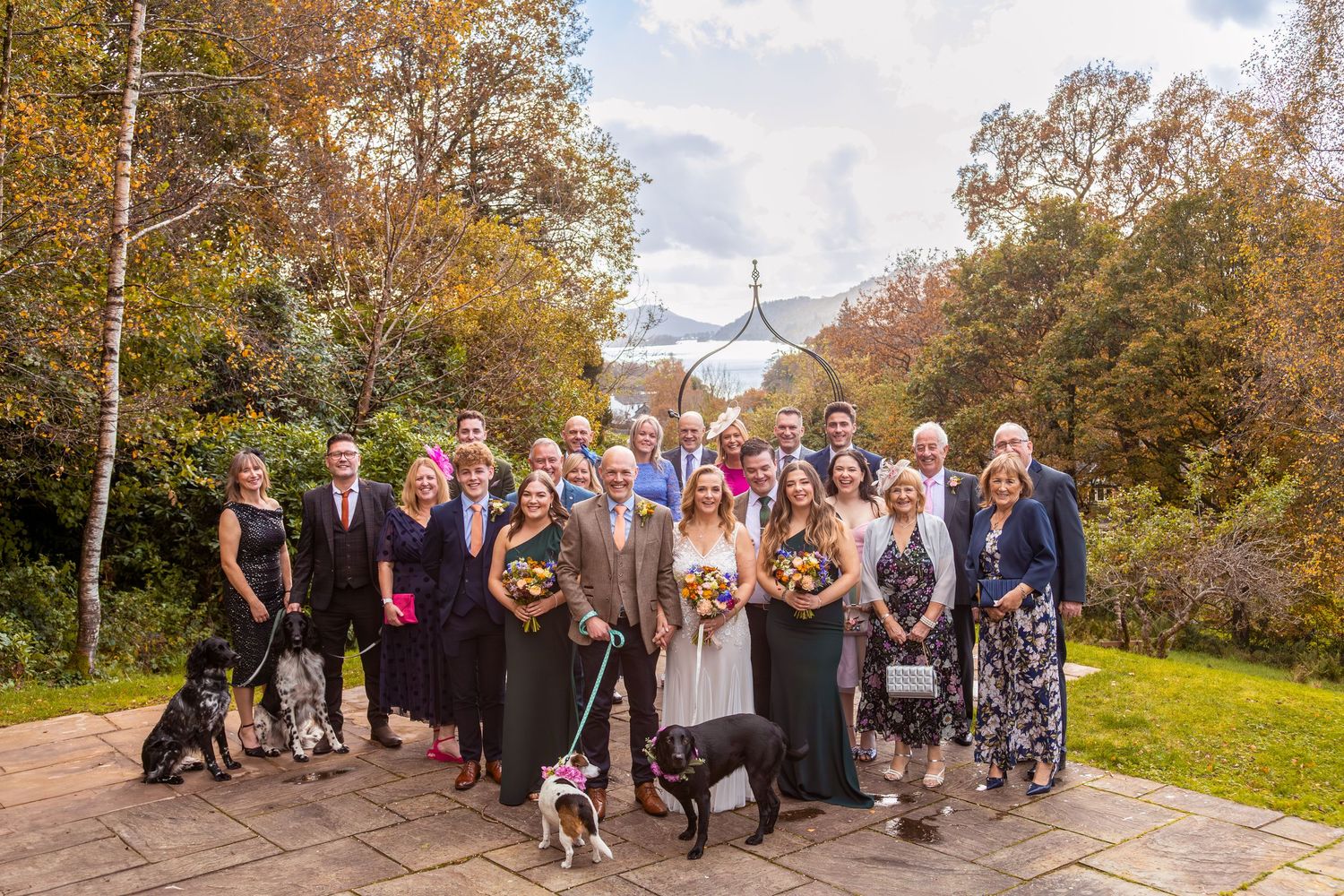 Wedding party group photo outdoors in autumn garden with dogs and colorful fall foliage backdrop.