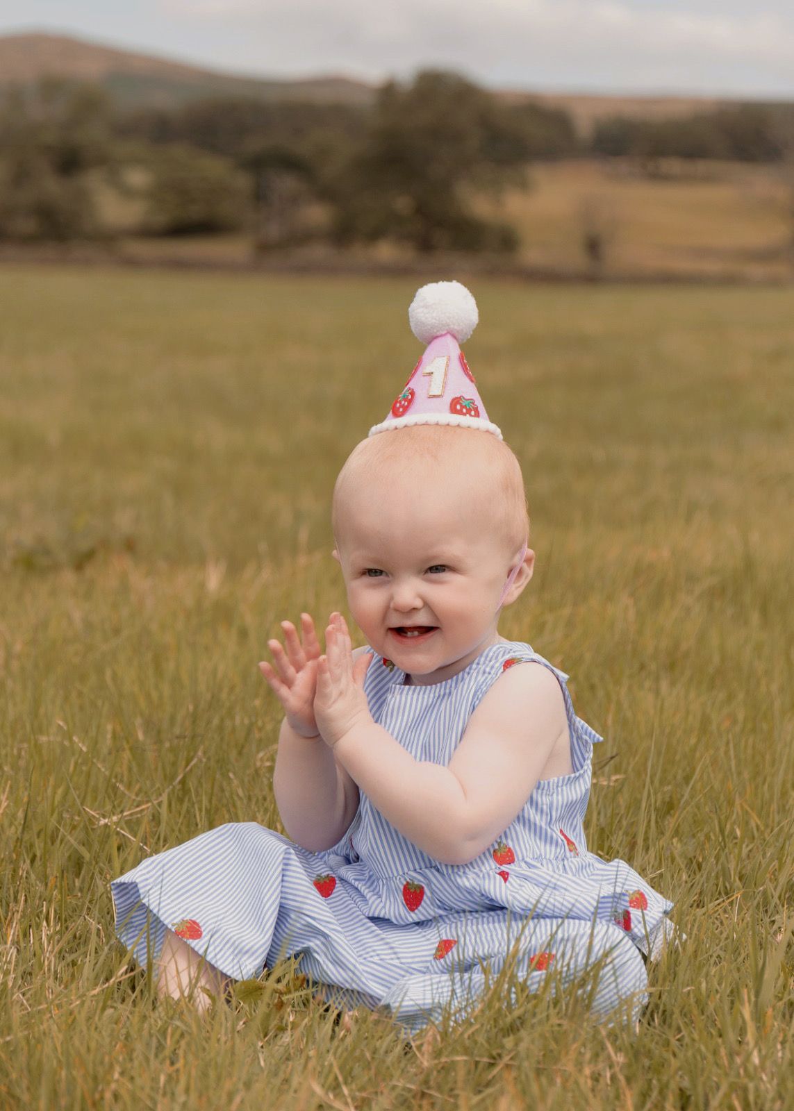 A joyful baby in a strawberry dress and birthday hat claps hands while sitting in a green field.