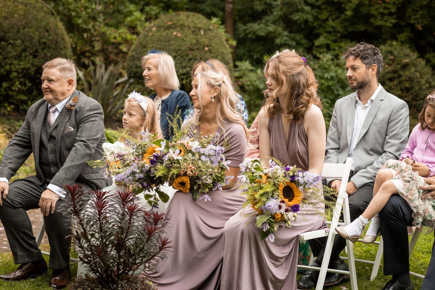 Bridesmaids in mauve dresses holding wildflower bouquets with sunflowers at outdoor ceremony.
