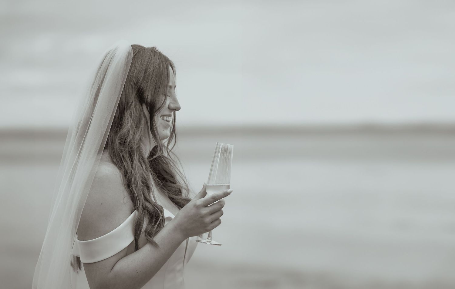 Elegant bride in white gown and veil holds champagne glass on misty beach in black and white photo.