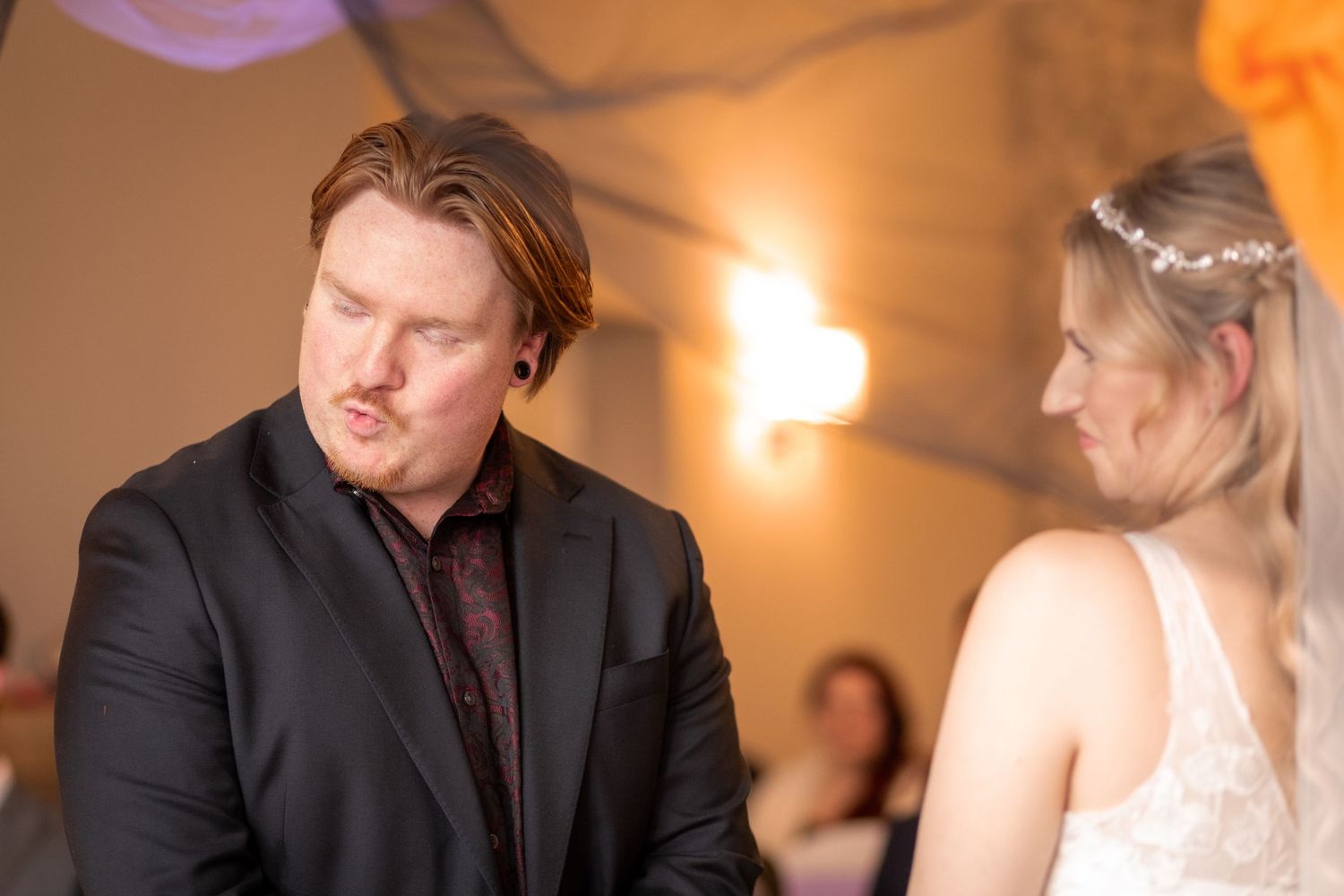 Groom in black suit exchanges emotional vows with bride in white lace dress during indoor wedding ceremony.