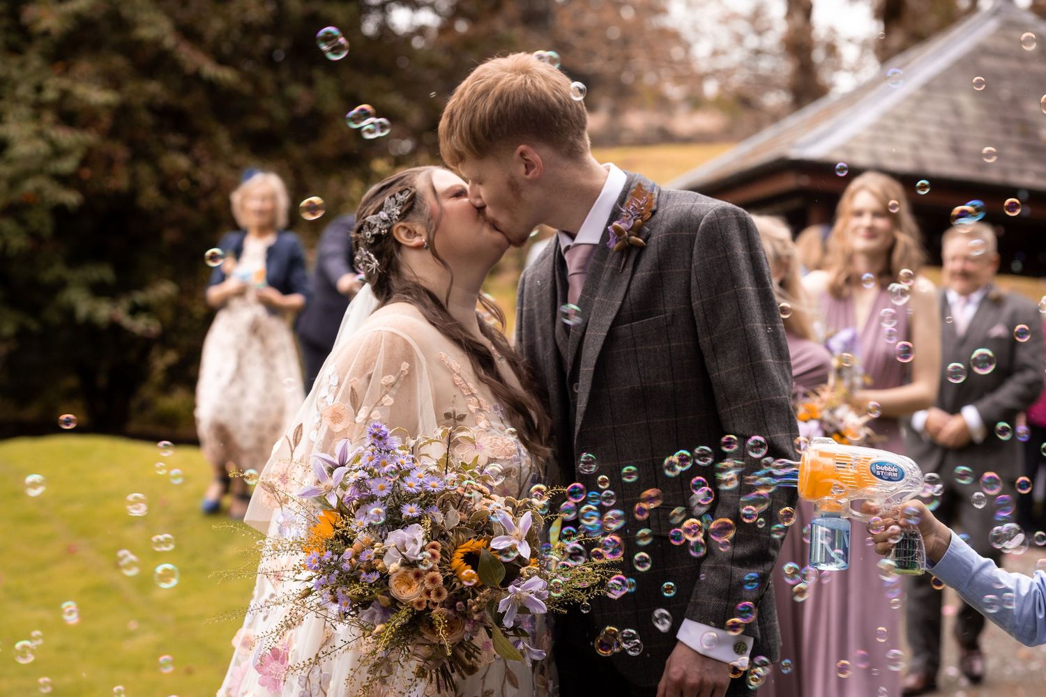 Bride and groom kissing surrounded by bubbles at outdoor wedding ceremony, bride holding wildflower bouquet.