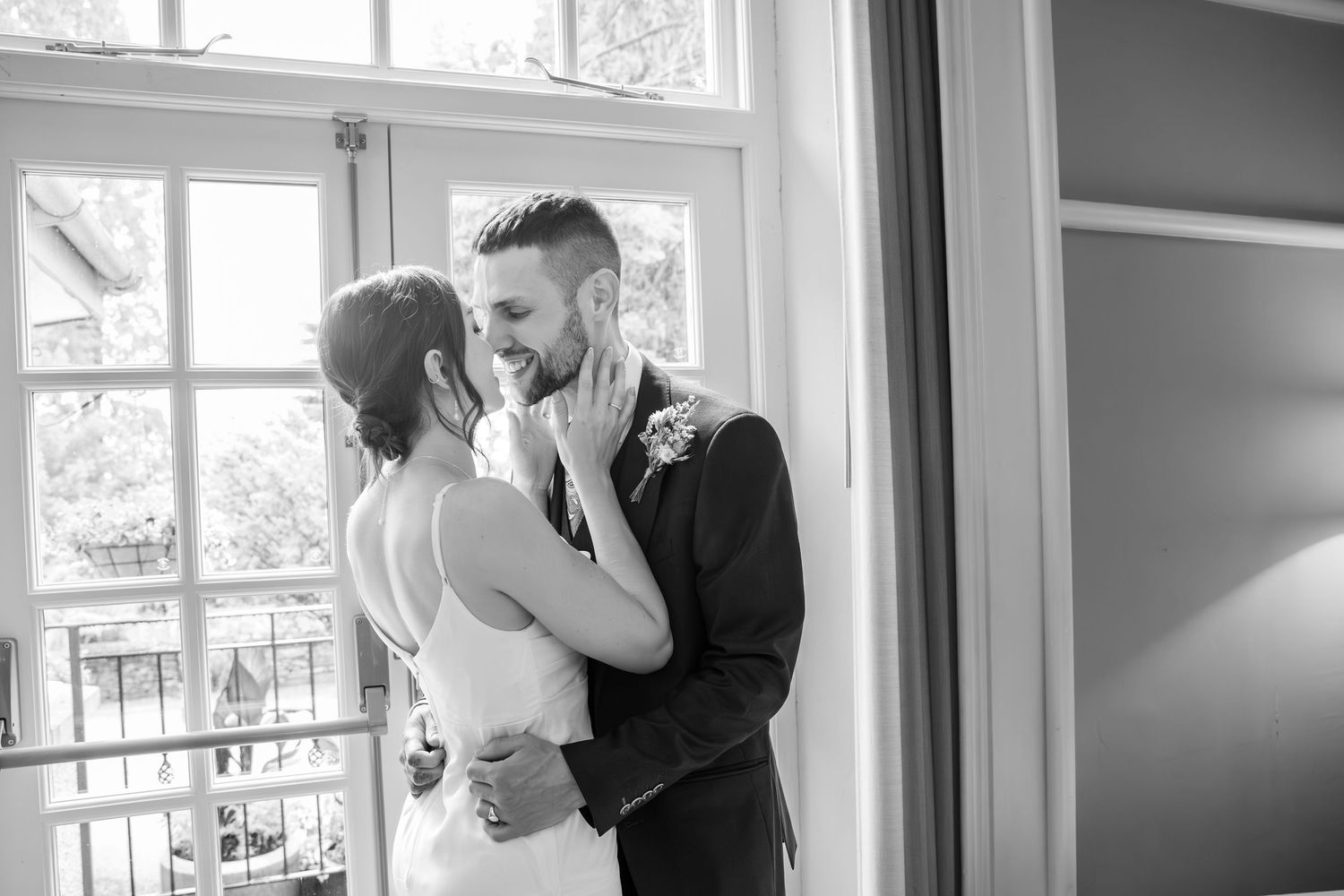 Black and white photo of a smiling bride and groom sharing a tender moment by a window on their wedding day.