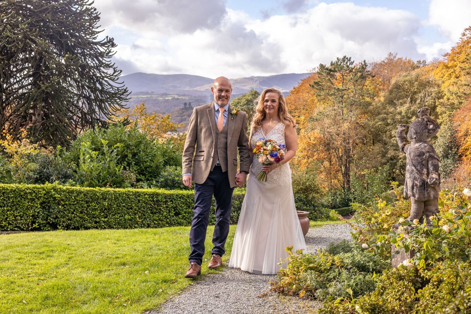 Bride and groom walking through autumn garden with colorful foliage and mountain backdrop.