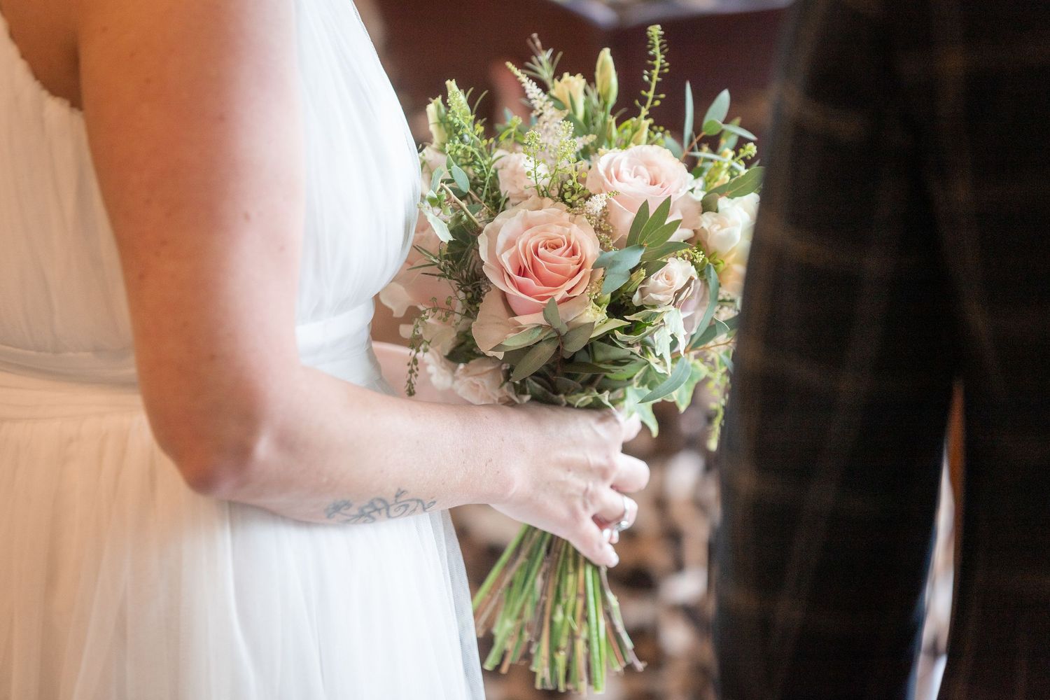 Bride holding a delicate pink rose and greenery wedding bouquet during a ceremony.