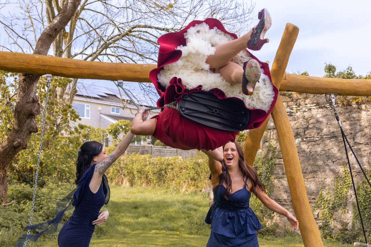 Bridesmaids laughing while playfully lifting Santa Claus upside down on outdoor playground equipment.