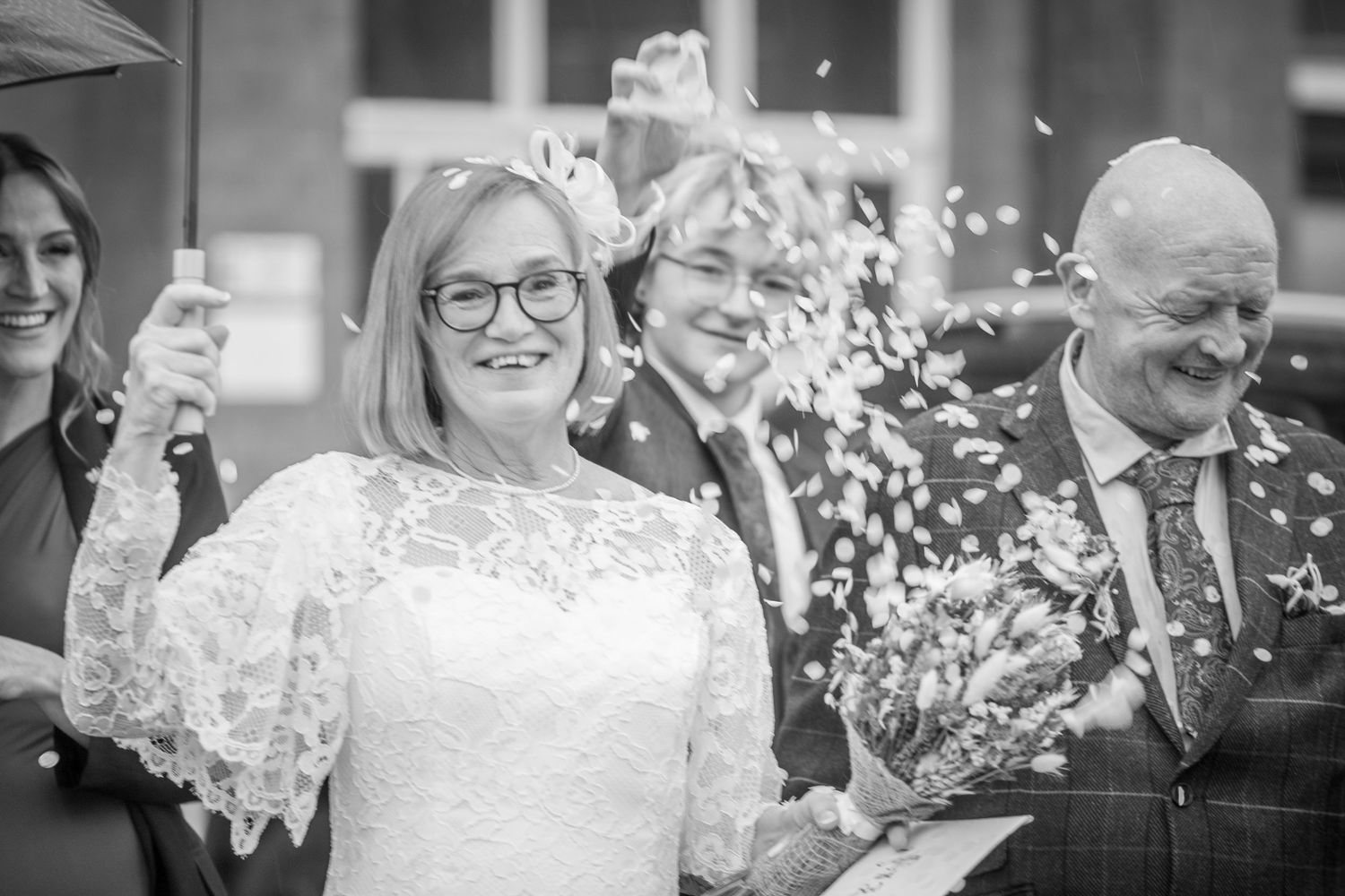Black and white photo of smiling bride with glasses enjoying confetti celebration after ceremony.