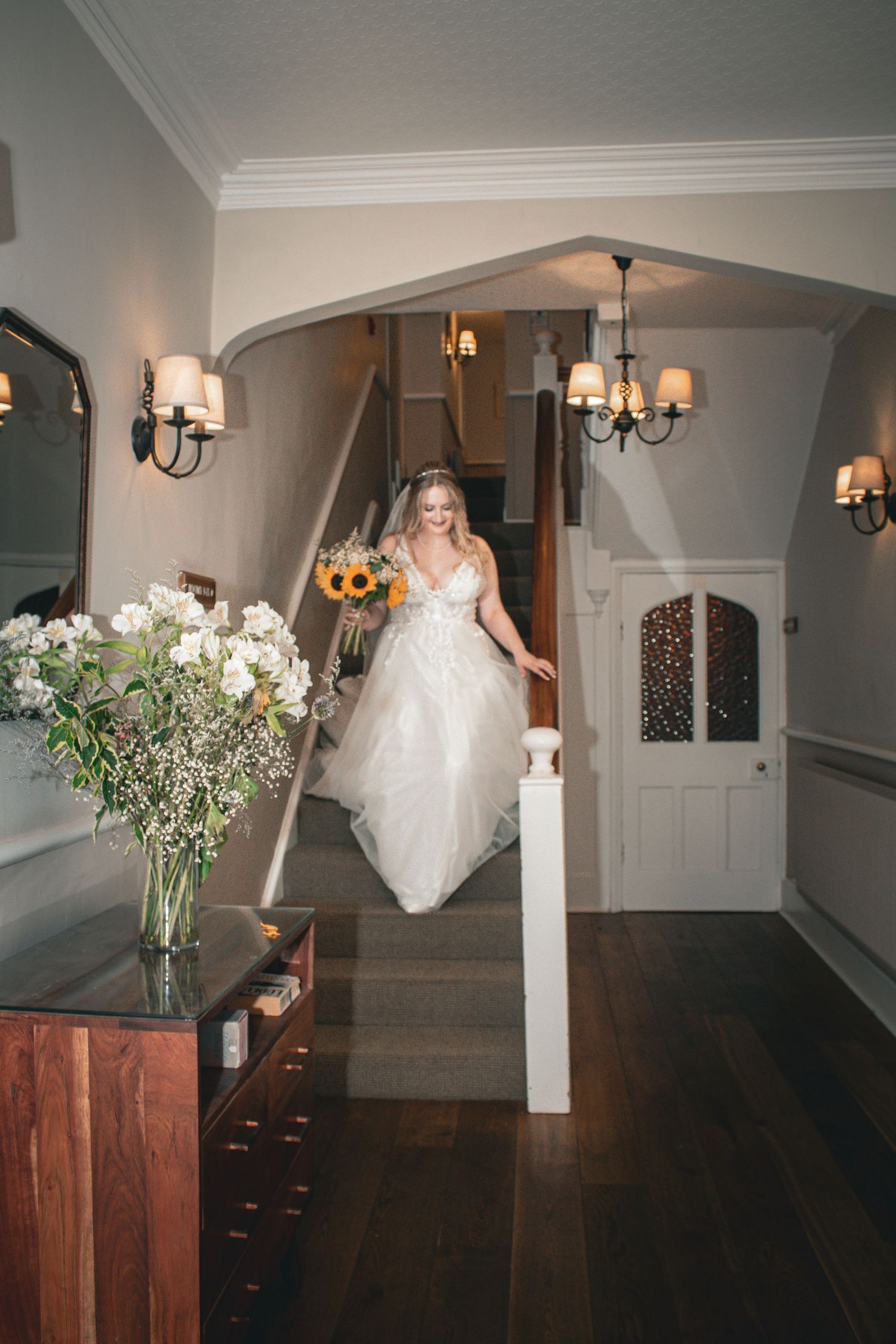 Elegant bride in lace gown descends staircase holding sunflower bouquet in a grand foyer.