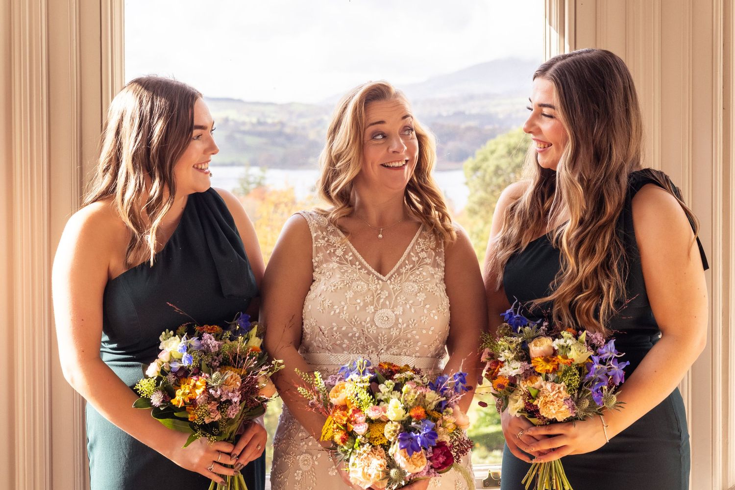 Bride with two bridesmaids holding colorful wildflower bouquets indoors.
