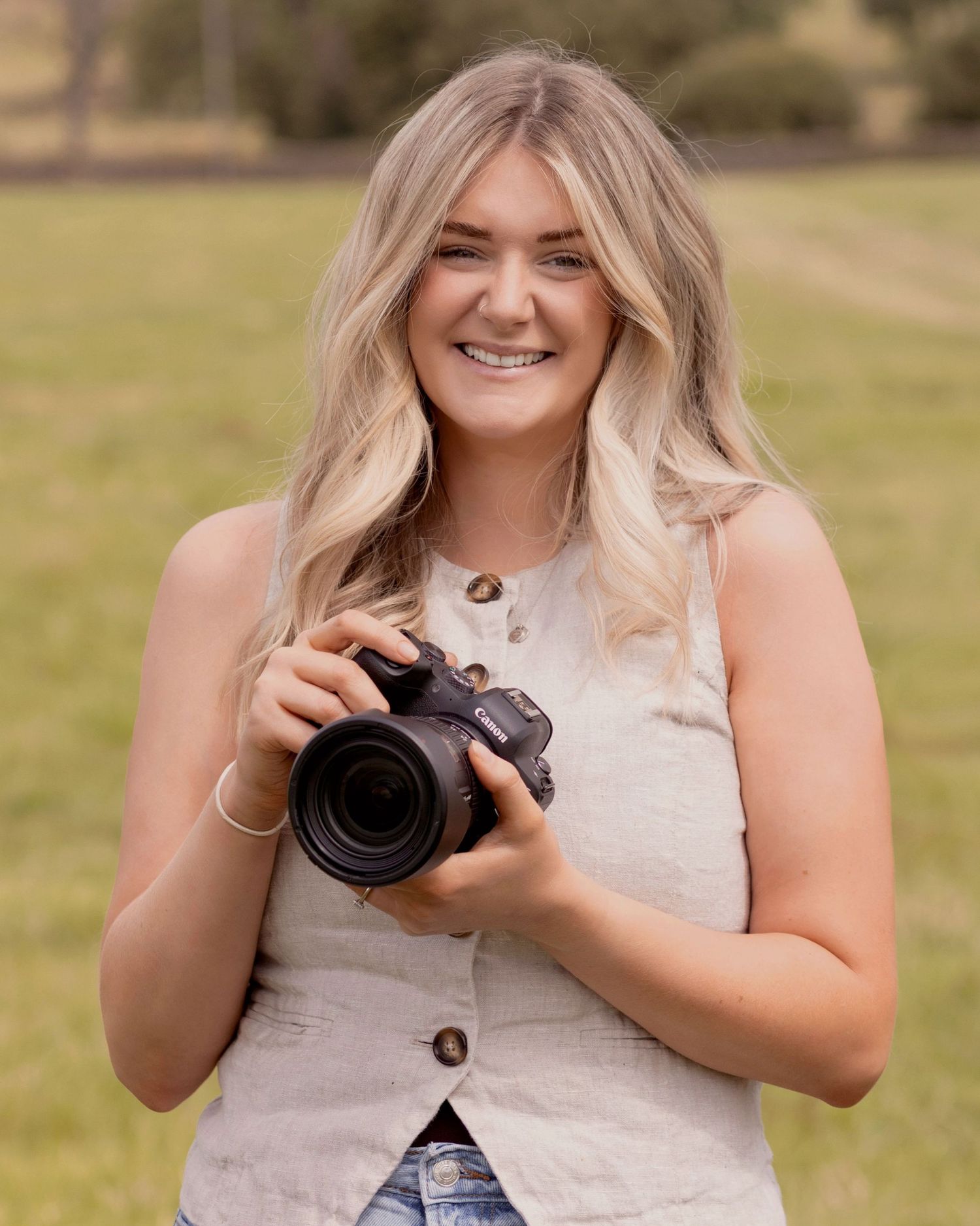 Young blonde woman smiling outdoors holding a DSLR camera, wearing a linen vest top in a green field.