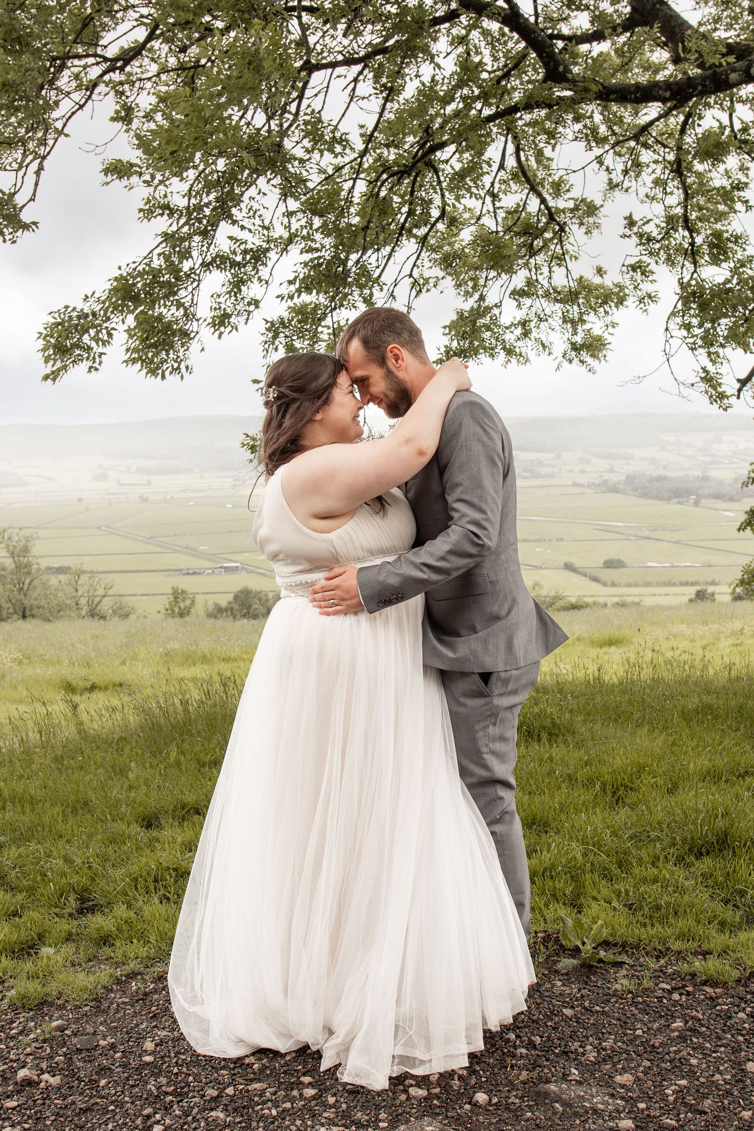 Bride and groom embracing under a tree with lush green countryside in the background on their wedding day.