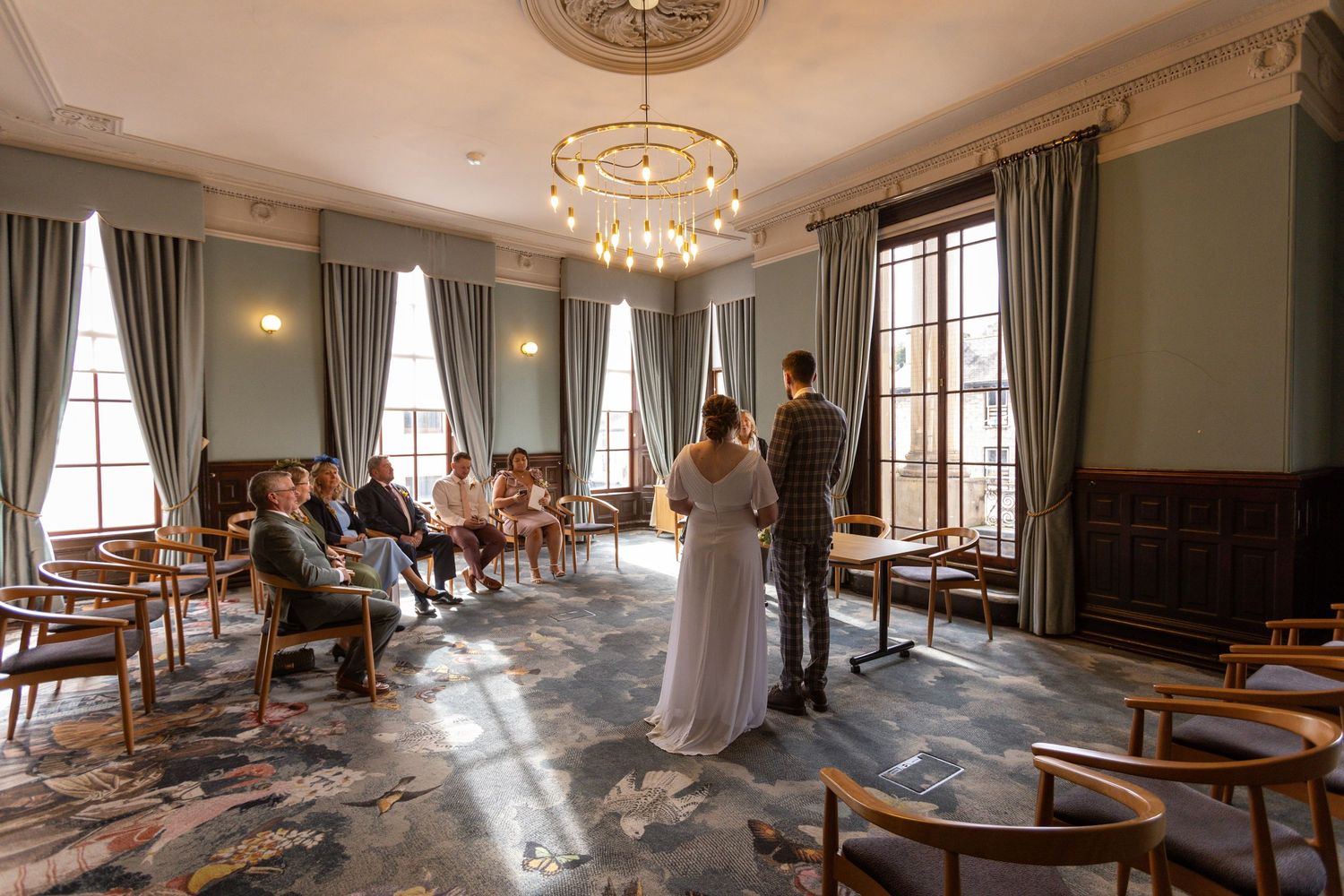 Intimate wedding ceremony in an elegant room with tall windows and a gold chandelier.