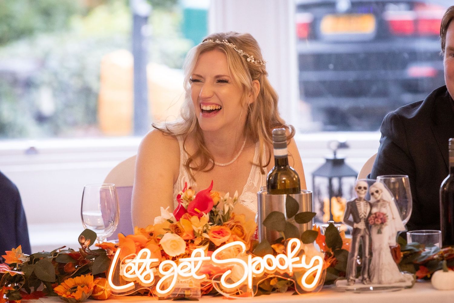 Laughing bride at wedding reception table with colorful floral arrangements and neon sign.