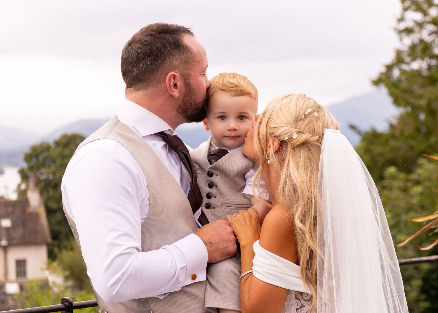 Bride and groom kissing their young son on cheeks on their wedding day with scenic greenery backdrop.
