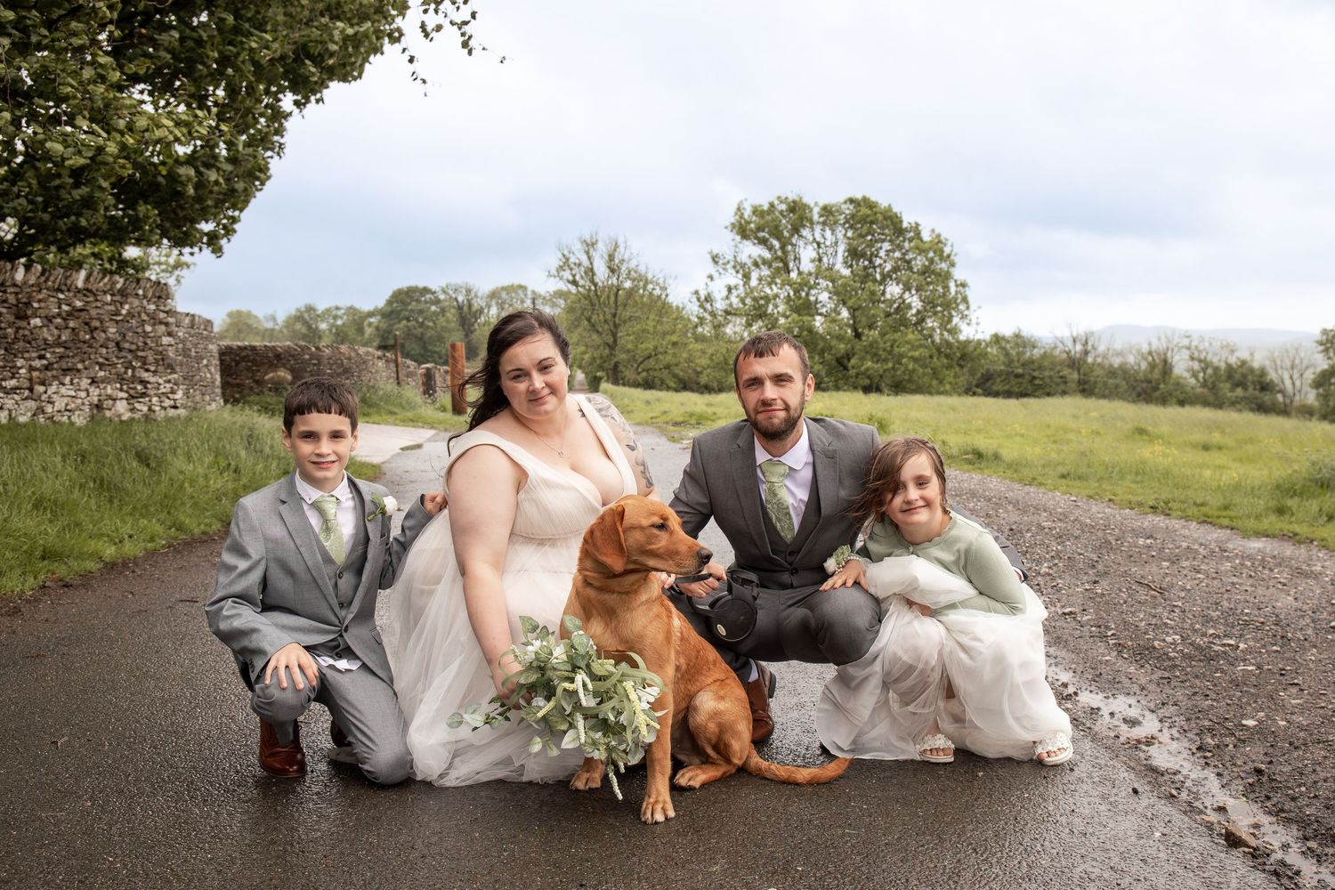 Wedding couple with two children and a golden dog posing on a country road surrounded by green fields.