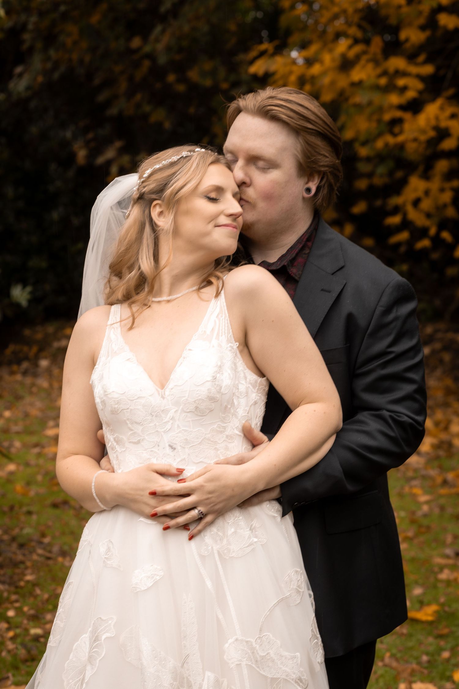 Bride and groom share a tender moment outdoors in autumn, groom kissing bride's cheek as she smiles in white lace gown.