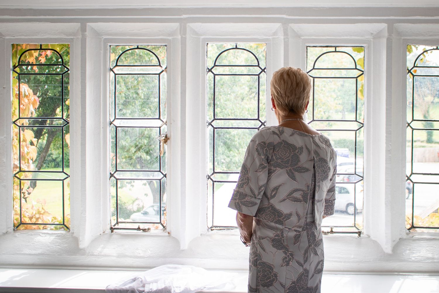 Woman in floral dress gazes through beautiful stained glass windows with garden views beyond.